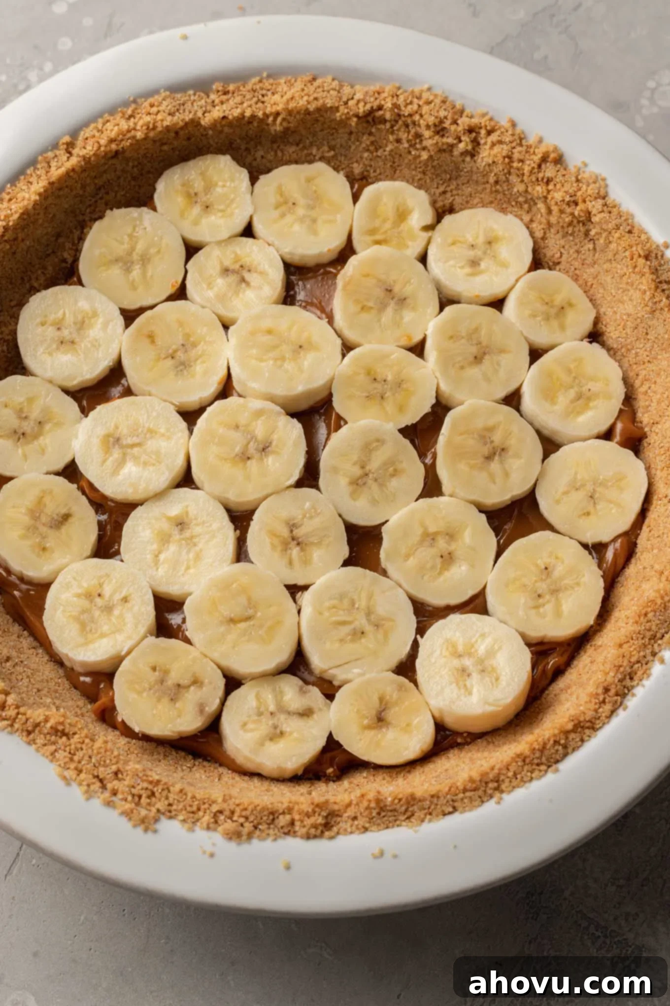 An overhead view of the banoffee pie in progress, showing the first layer of dulce de leche topped with neatly arranged banana slices in the graham cracker crust. 