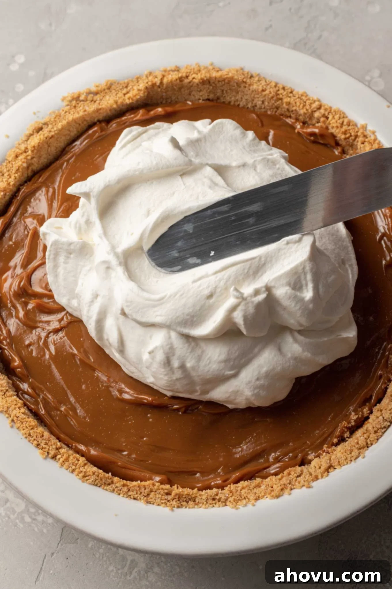 Creamy whipped cream being spread generously over the dulce de leche and banana layers in a pie dish, creating a smooth top. 