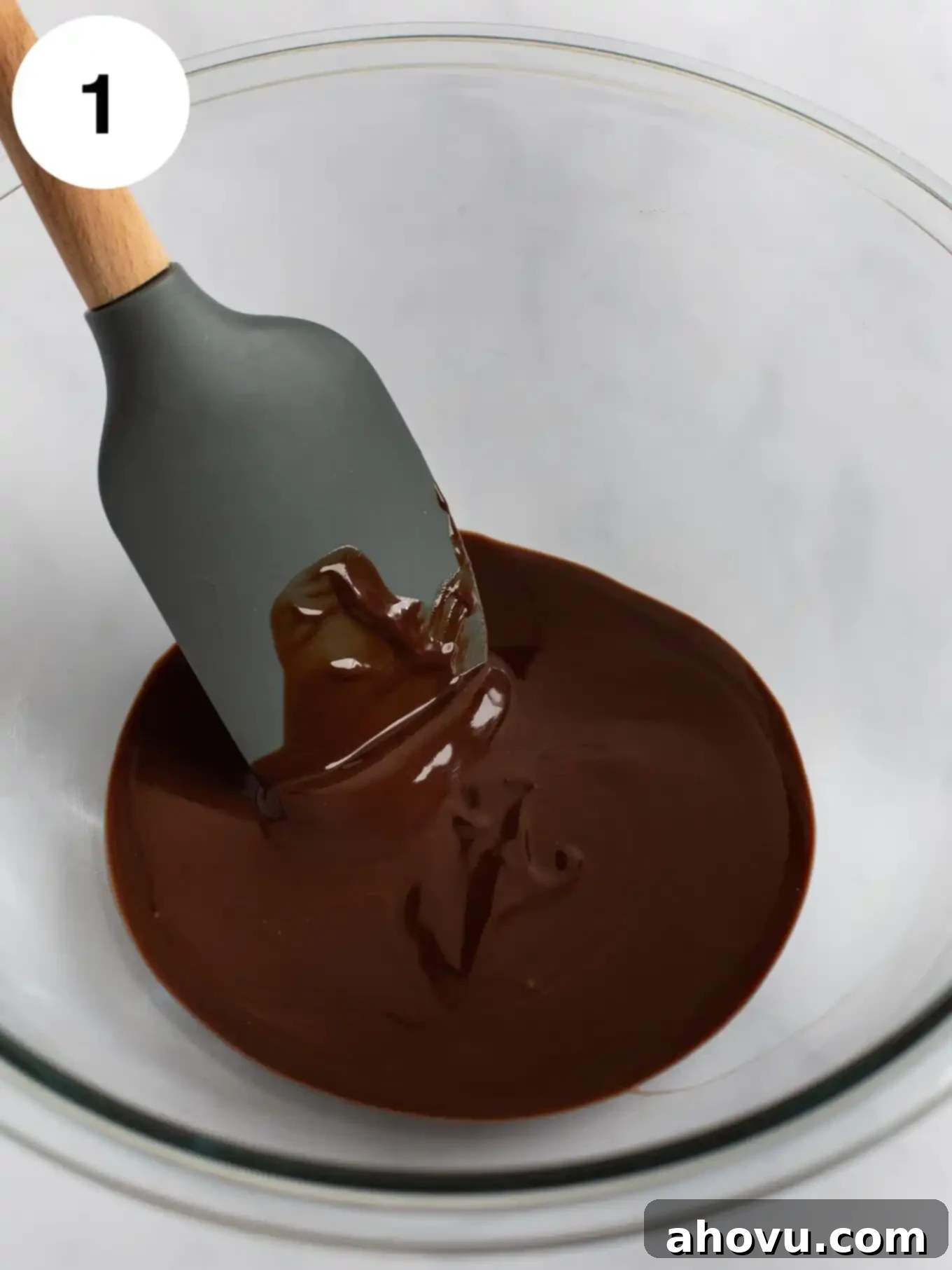 Melted semi-sweet chocolate and unsalted butter are mixed together in a clear glass mixing bowl, with a rubber spatula resting inside.