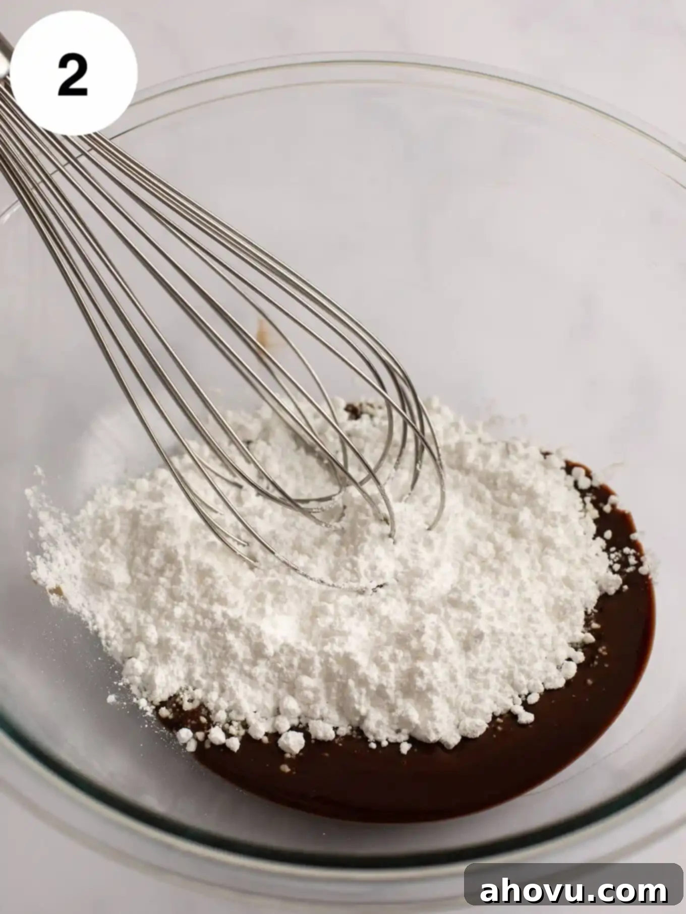 A stream of white powdered sugar is being gently added to the warm, melted butter and chocolate mixture in a glass bowl.