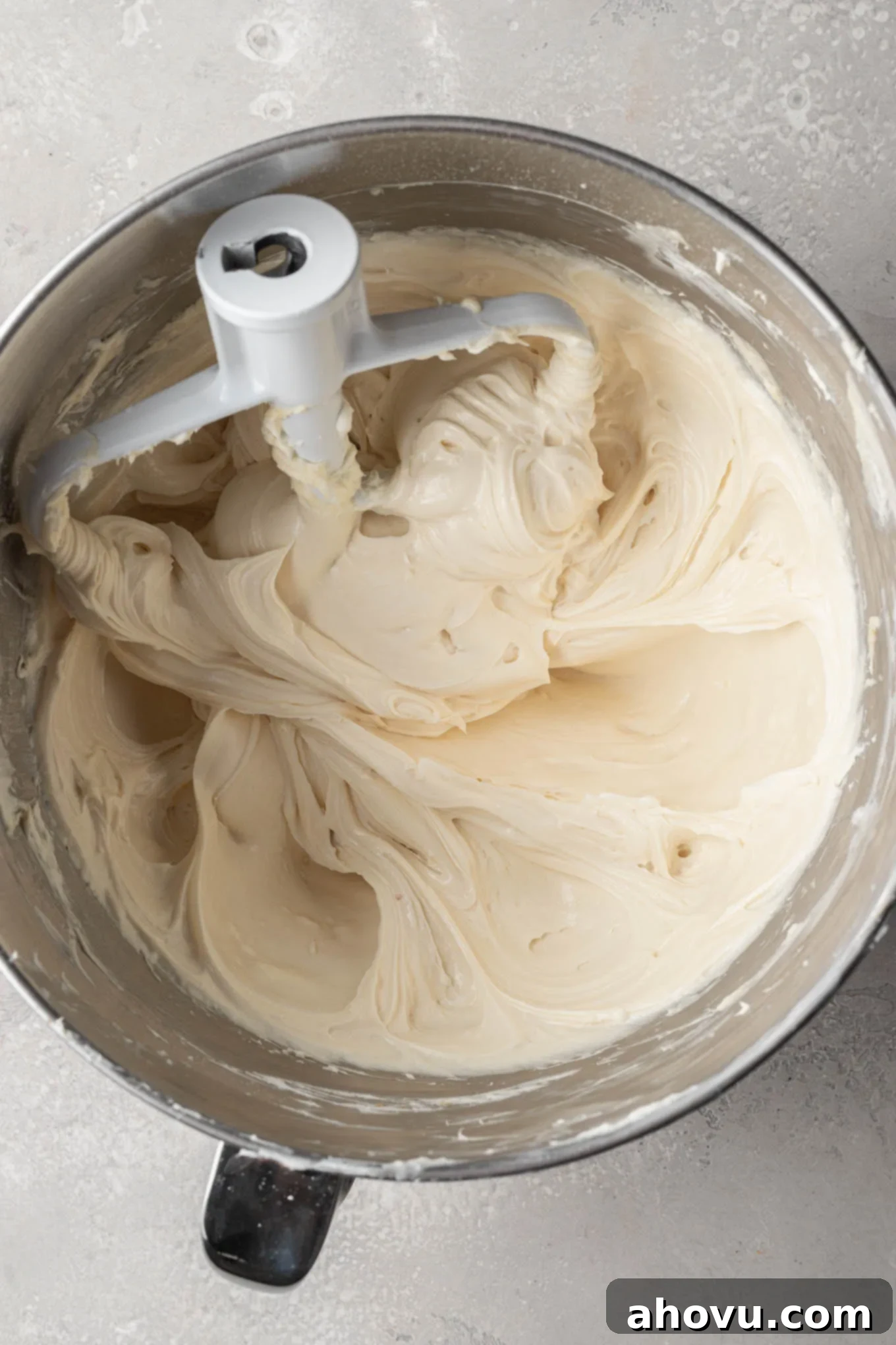An overhead view of softened cream cheese being beaten to a smooth consistency in the bowl of a stand mixer with a paddle attachment.