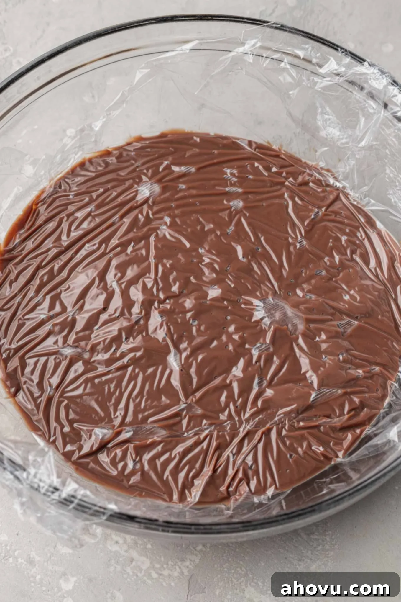An overhead view of homemade chocolate pudding in a glass mixing bowl, with plastic wrap pressed directly on top to prevent a skin from forming.