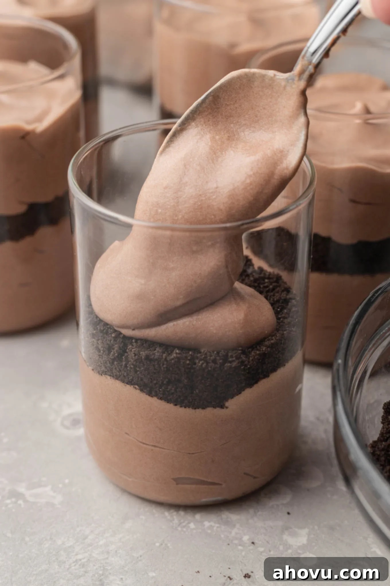A process shot showing creamy chocolate pudding being spooned on top of a layer of crushed Oreo crumbs in a clear cup.