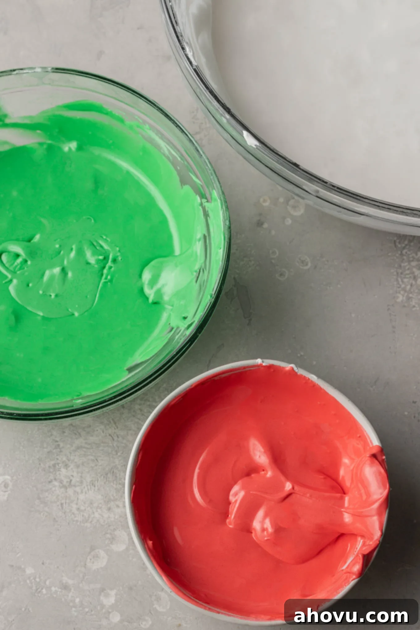 An overhead view of three separate mixing bowls, neatly filled with vibrant green, festive red, and pure white royal icing, ready for holiday cookie decoration.