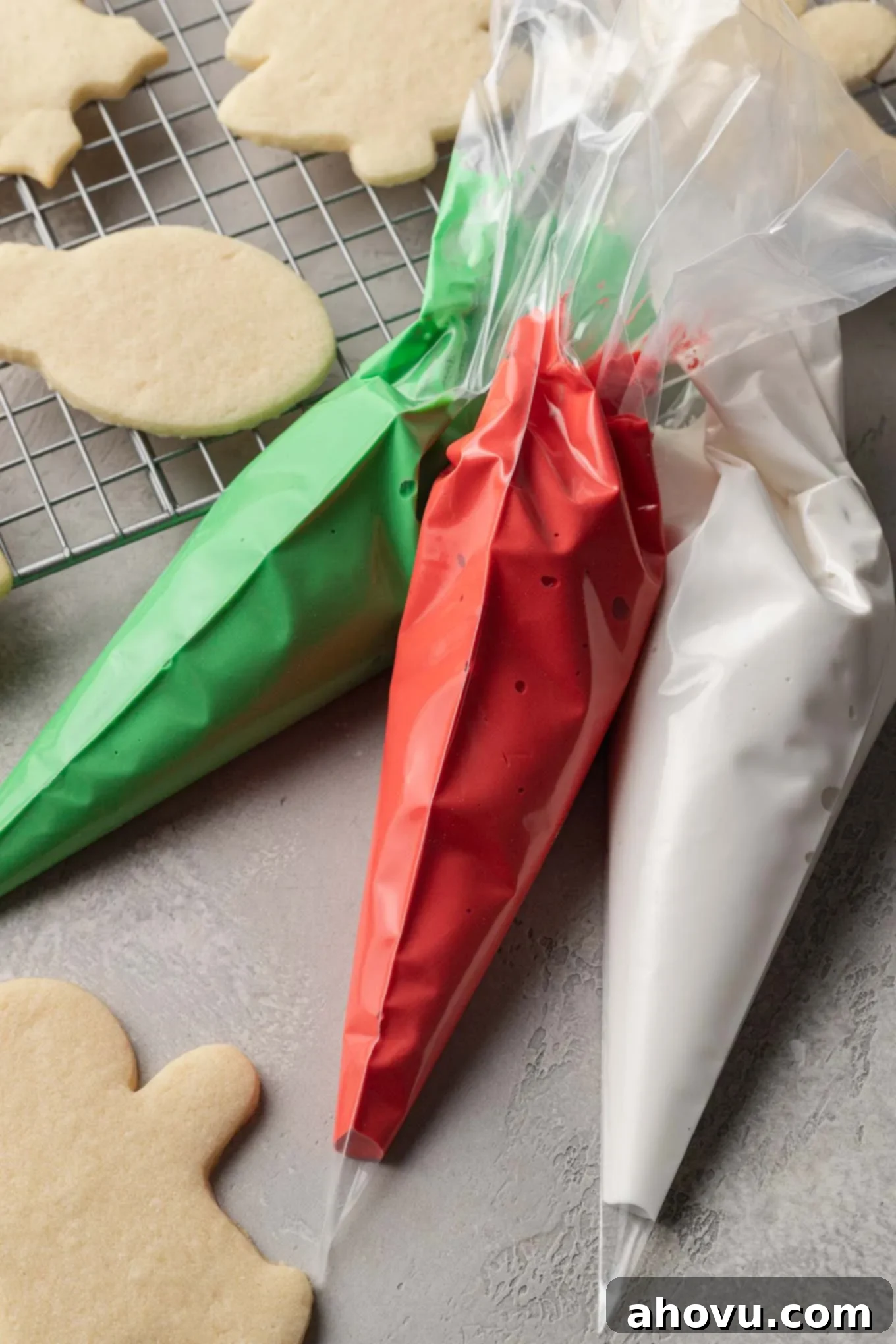 A festive tableau featuring three piping bags, brimming with red, green, and white royal icing, arranged beside a cooling rack adorned with freshly baked sugar cookies.
