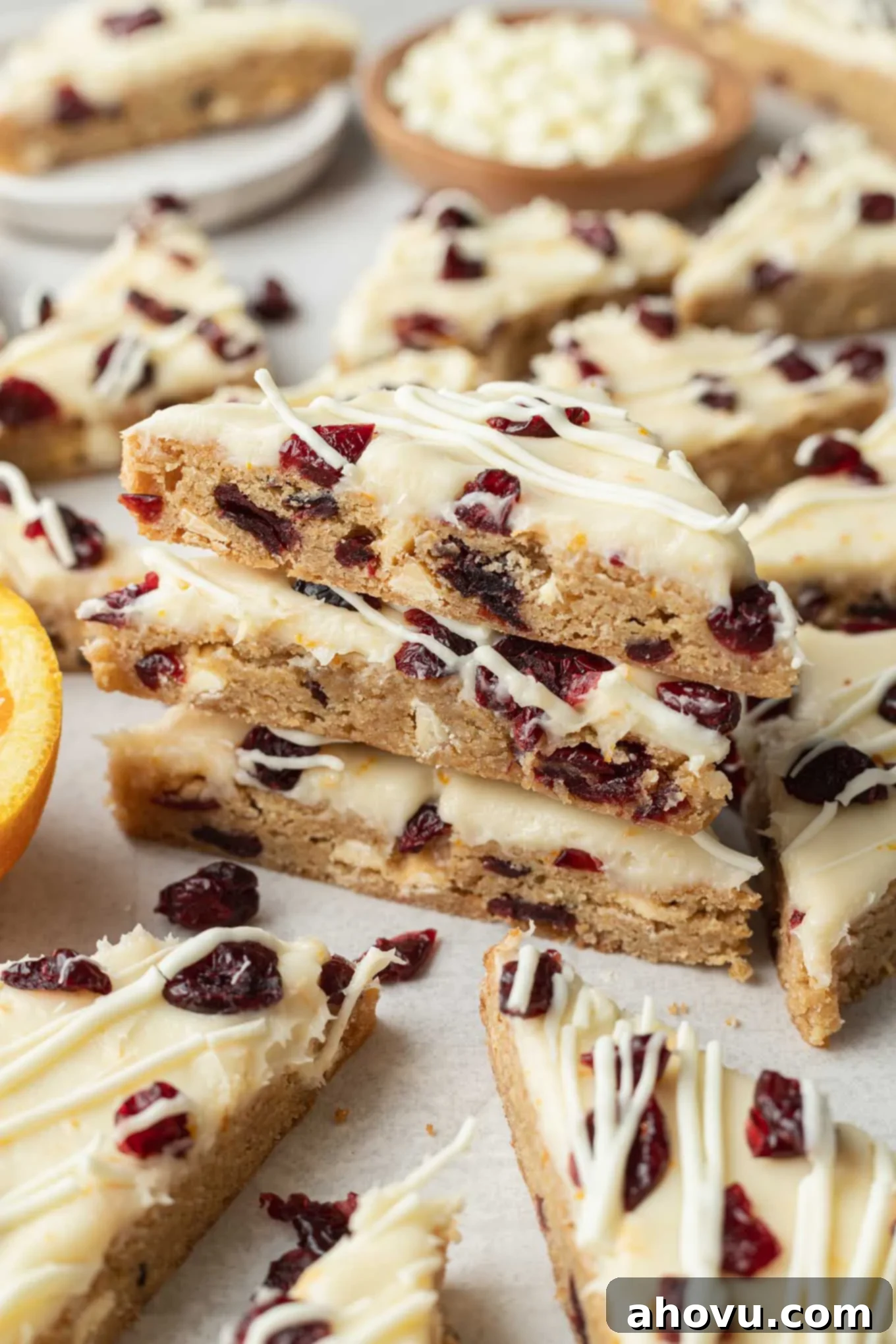 A stack of three homemade cranberry bliss bars, showing the blondie base, orange cream cheese frosting, and white chocolate drizzle, surrounded by additional bars ready to be enjoyed.
