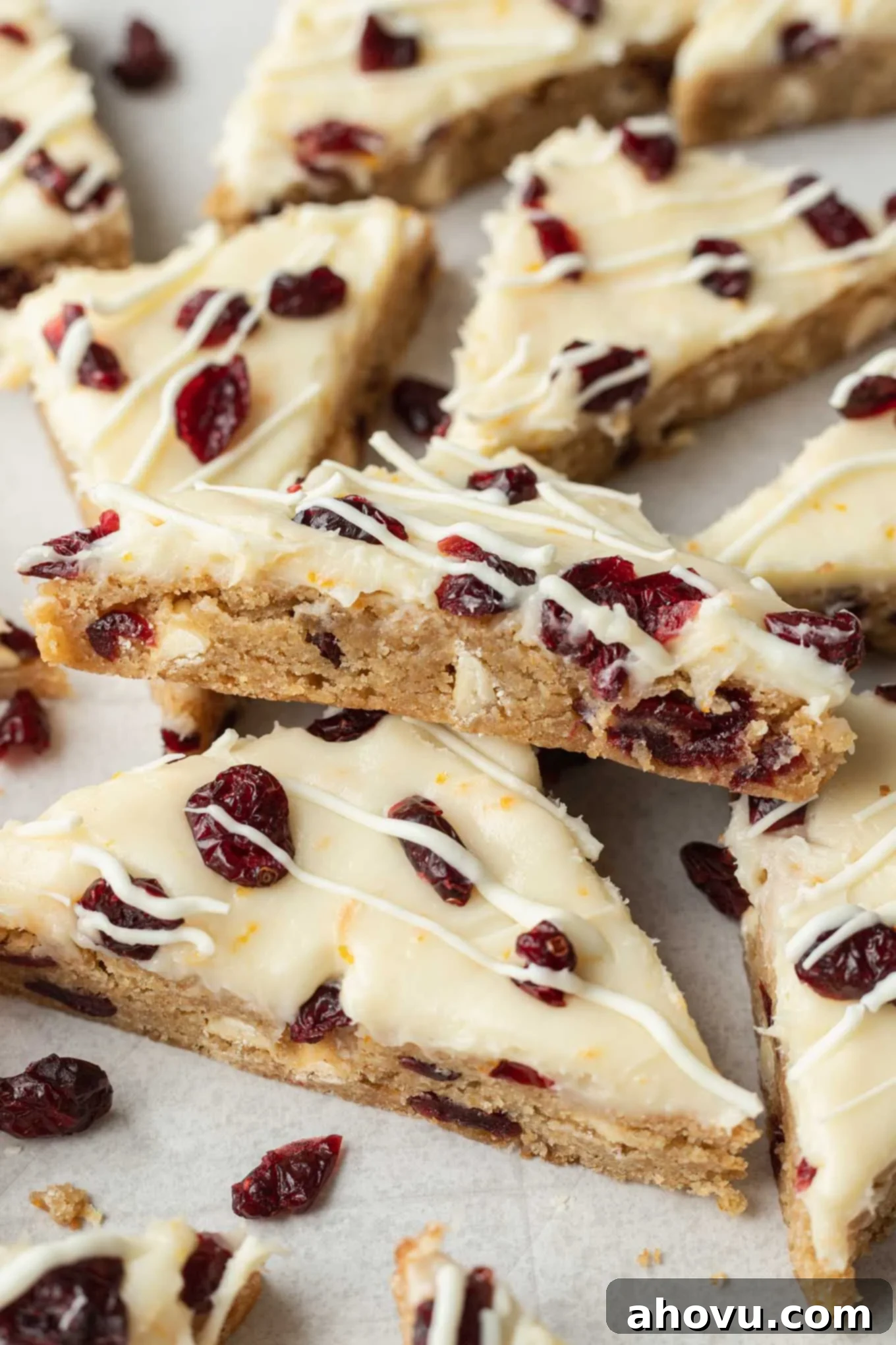 A close-up side view of a single cranberry bliss bar, showcasing its distinct layers: the chewy blondie base, the thick layer of orange cream cheese frosting, and the decorative white chocolate drizzle with dried cranberries, resting on another bar.