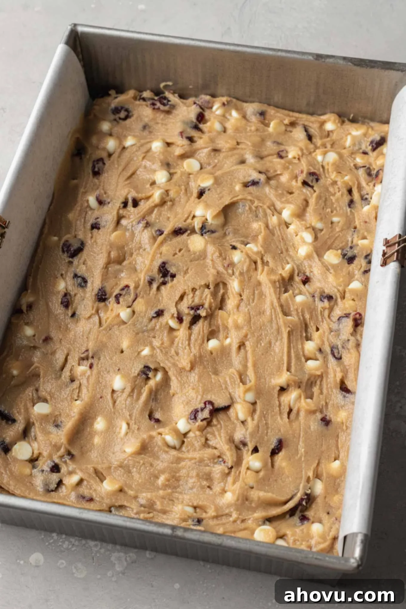 An overhead view of the raw cranberry bliss bar batter evenly spread in a parchment paper-lined 9x13 inch baking dish, showing the vibrant cranberries and white chocolate chips nestled within, before baking.