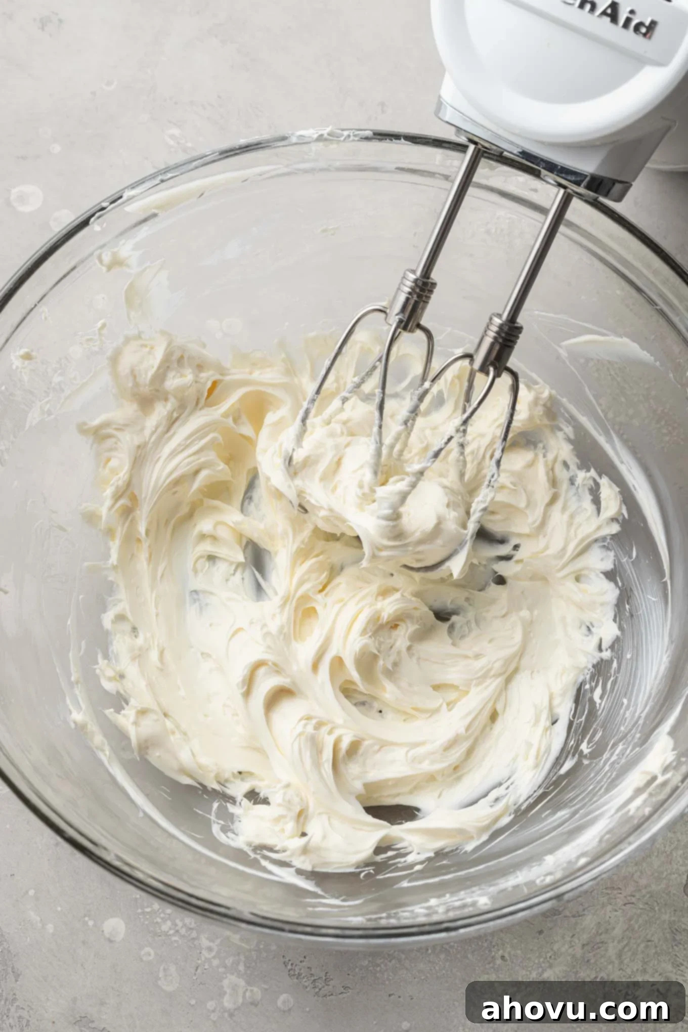 An overhead view of softened brick-style cream cheese being beaten until smooth in a glass mixing bowl with a handheld electric mixer, demonstrating the initial step for making the frosting.