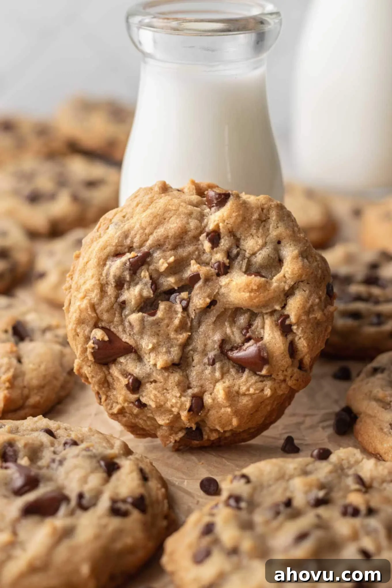A large bakery style chocolate chip cookie leaning against a tall glass of milk. More cookies are surrounding it.