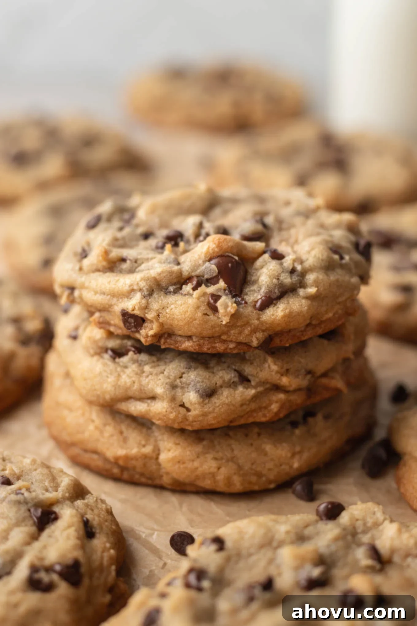 A stack of three bakery style chocolate chips cookies on a piece of brown parchment paper. More cookies are surrounding the stack of cookies.