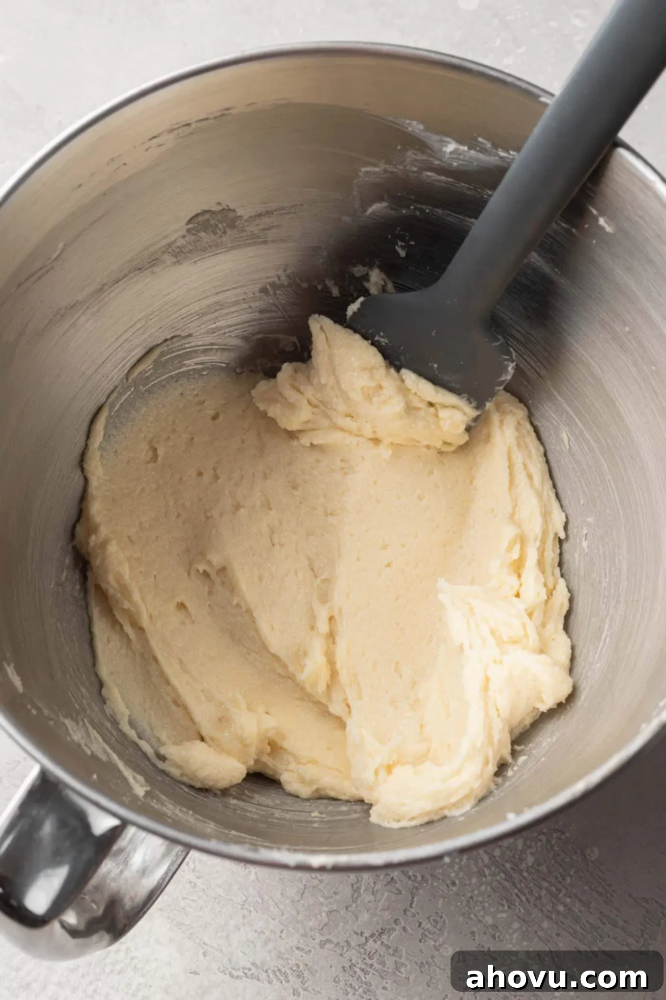 An overhead view of the wet ingredients for sugar cookies, including butter, sugar, egg, and vanilla, combined in a mixing bowl, with a rubber spatula nearby.