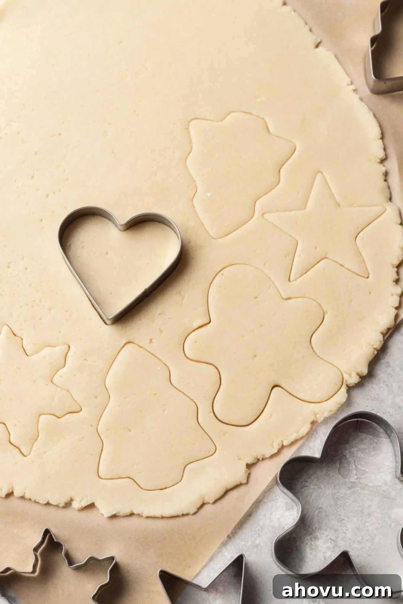 An overhead view of sugar cookie dough being cut into festive shapes using cookie cutters on parchment paper.