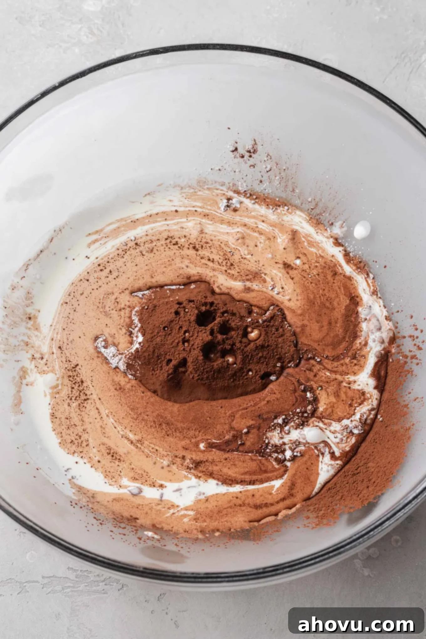 A glass mixing bowl with heavy cream, powdered sugar, cocoa powder, and vanilla extract.