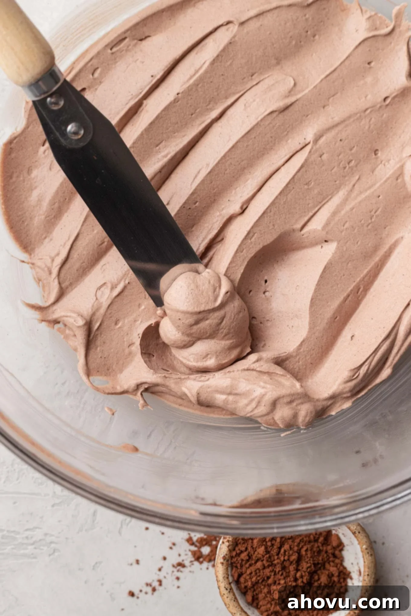 A glass mixing bowl filled with the finished chocolate whipped cream. An angled spatula is resting inside of the bowl.