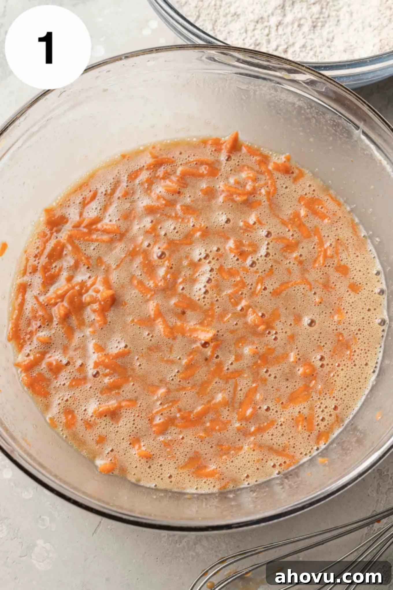 An overhead view of the wet ingredients for the carrot cake, including oil, sugars, sour cream, eggs, and vanilla, in a clear glass mixing bowl.