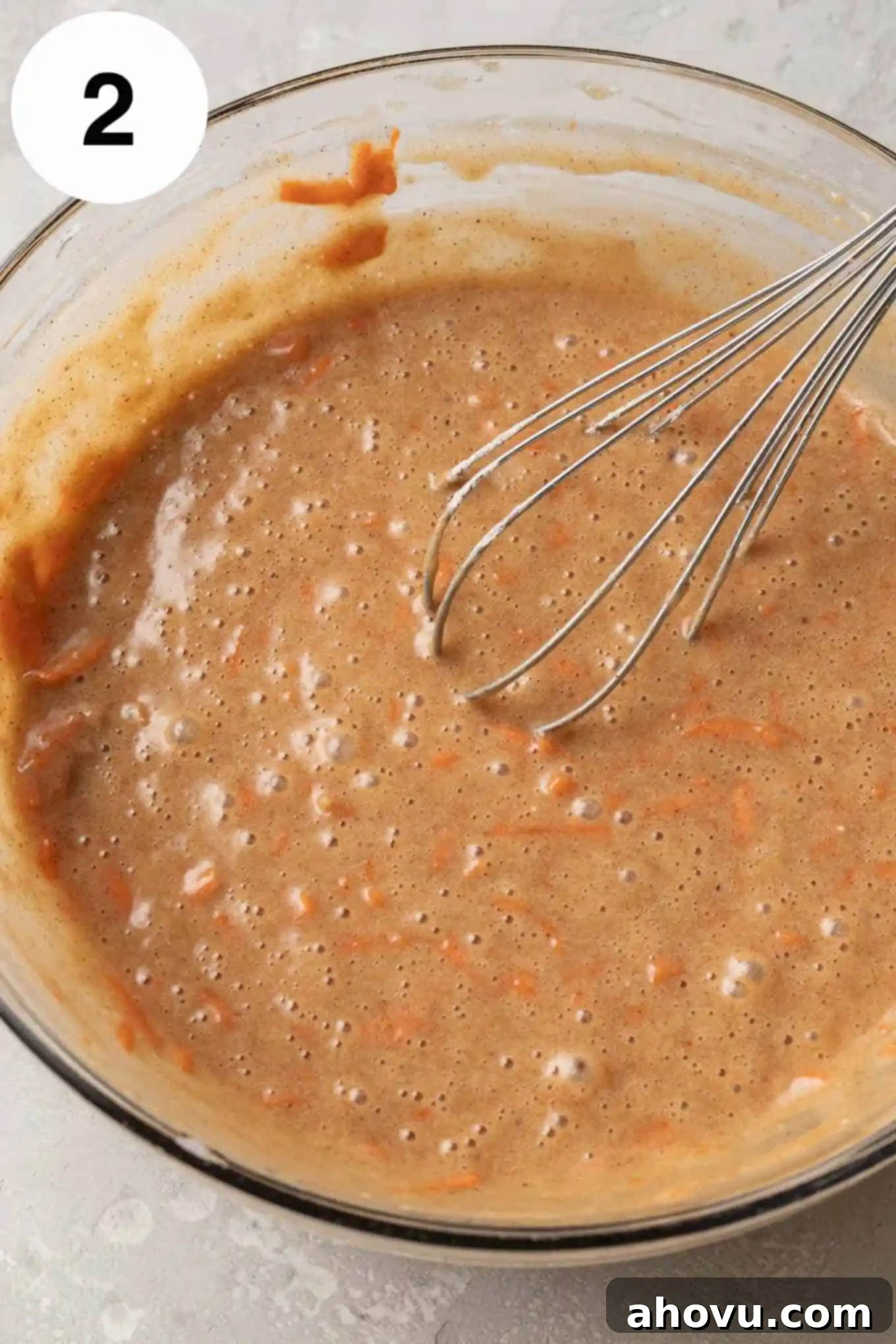 An overhead view of finished carrot cake batter in a glass mixing bowl with a whisk resting inside, showing the flecks of grated carrot.
