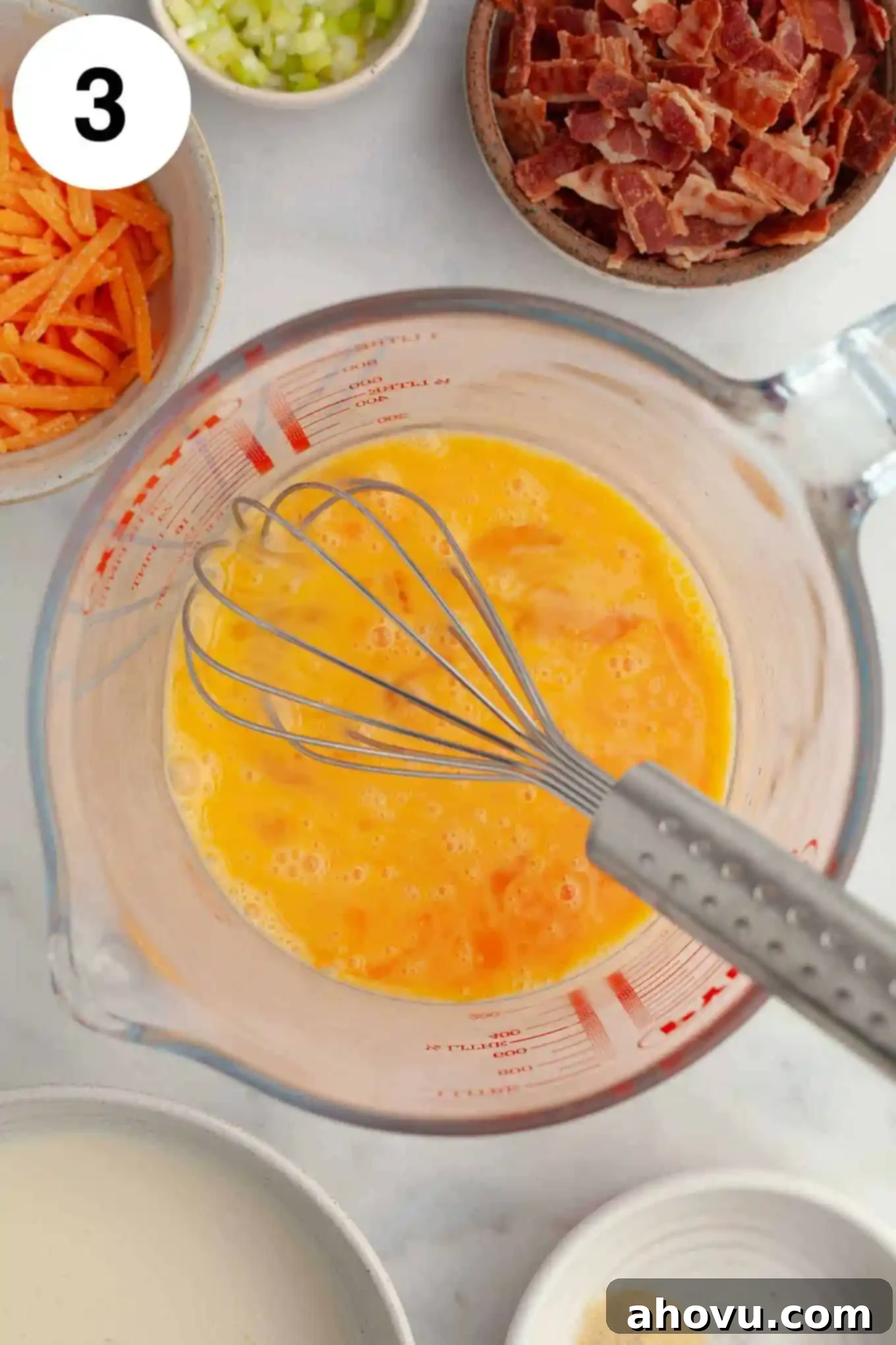 An overhead view of lightly beaten eggs in a glass measuring cup, with bowls of quiche mix-ins like bacon and cheese visible nearby.