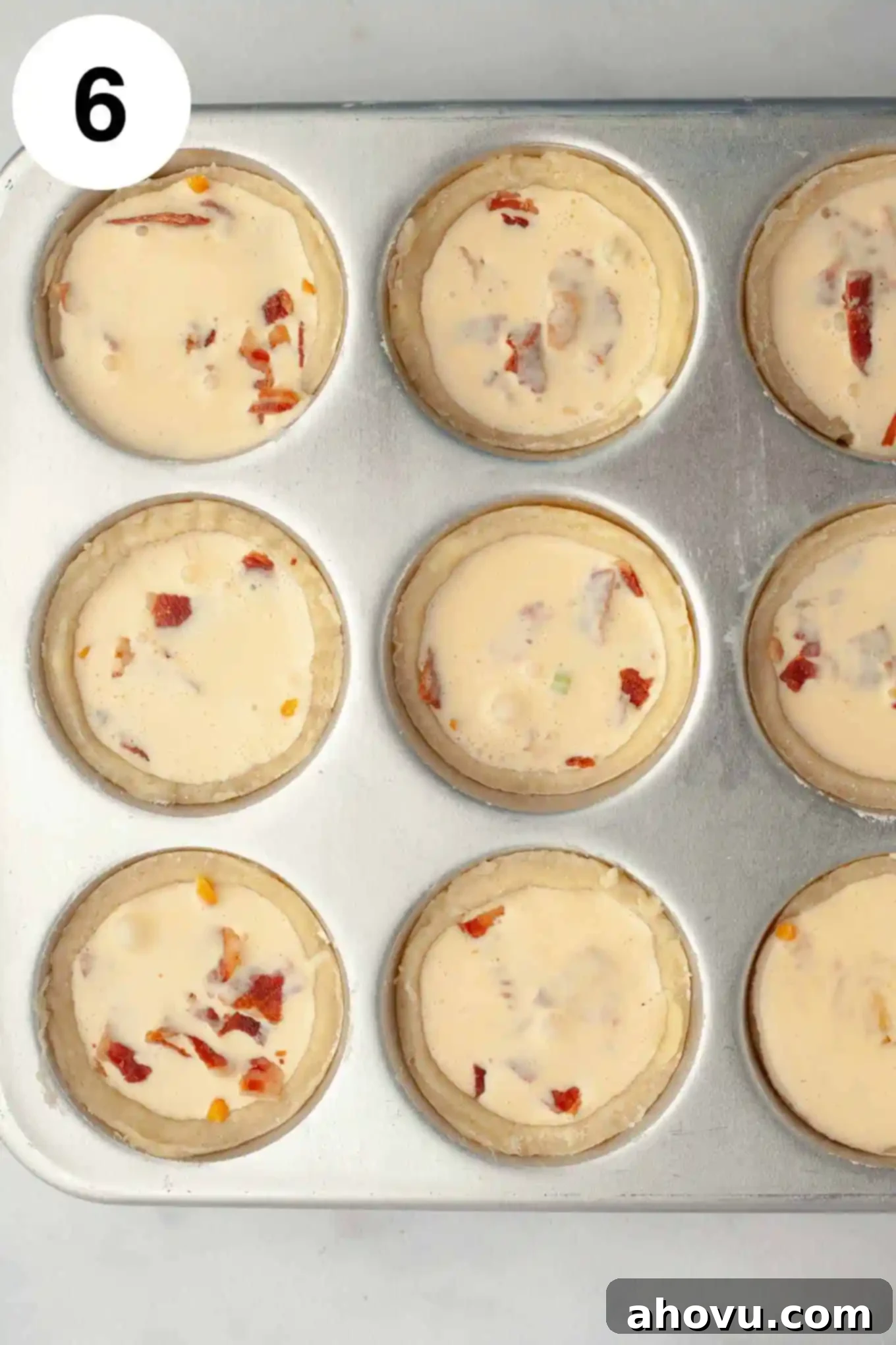 An overhead view of unbaked, filled mini quiches in a muffin pan, ready for the oven with their savory mix-ins and creamy egg filling.