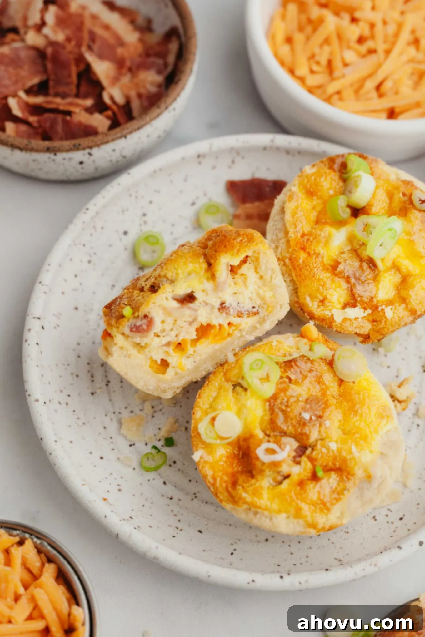An overhead view of golden-brown mini quiches arranged on a white plate. One quiche has been perfectly cut in half, revealing its creamy filling and savory mix-ins.