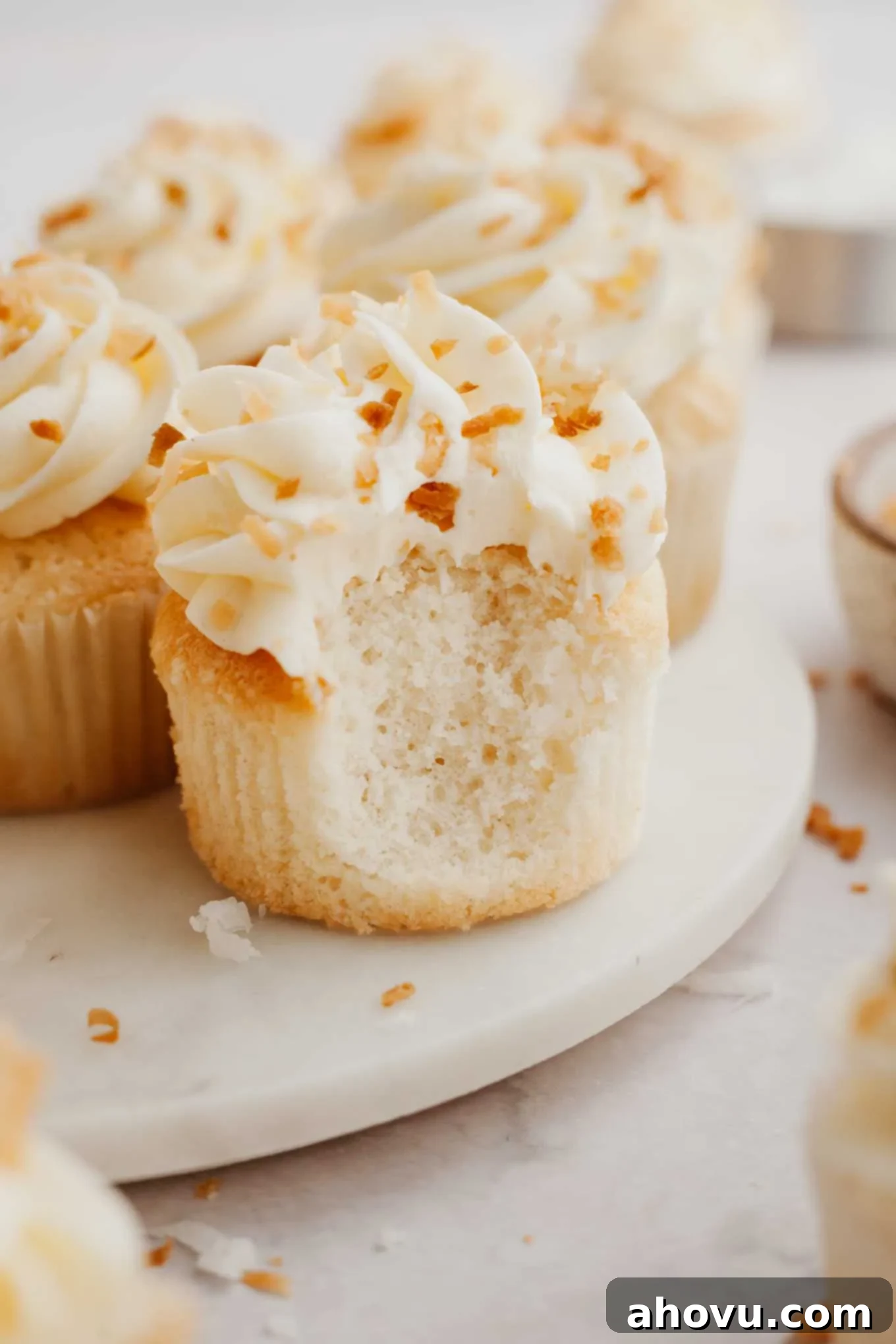 A perfectly frosted coconut cupcake with a bite taken out, revealing its fluffy interior, with more cupcakes in the background.