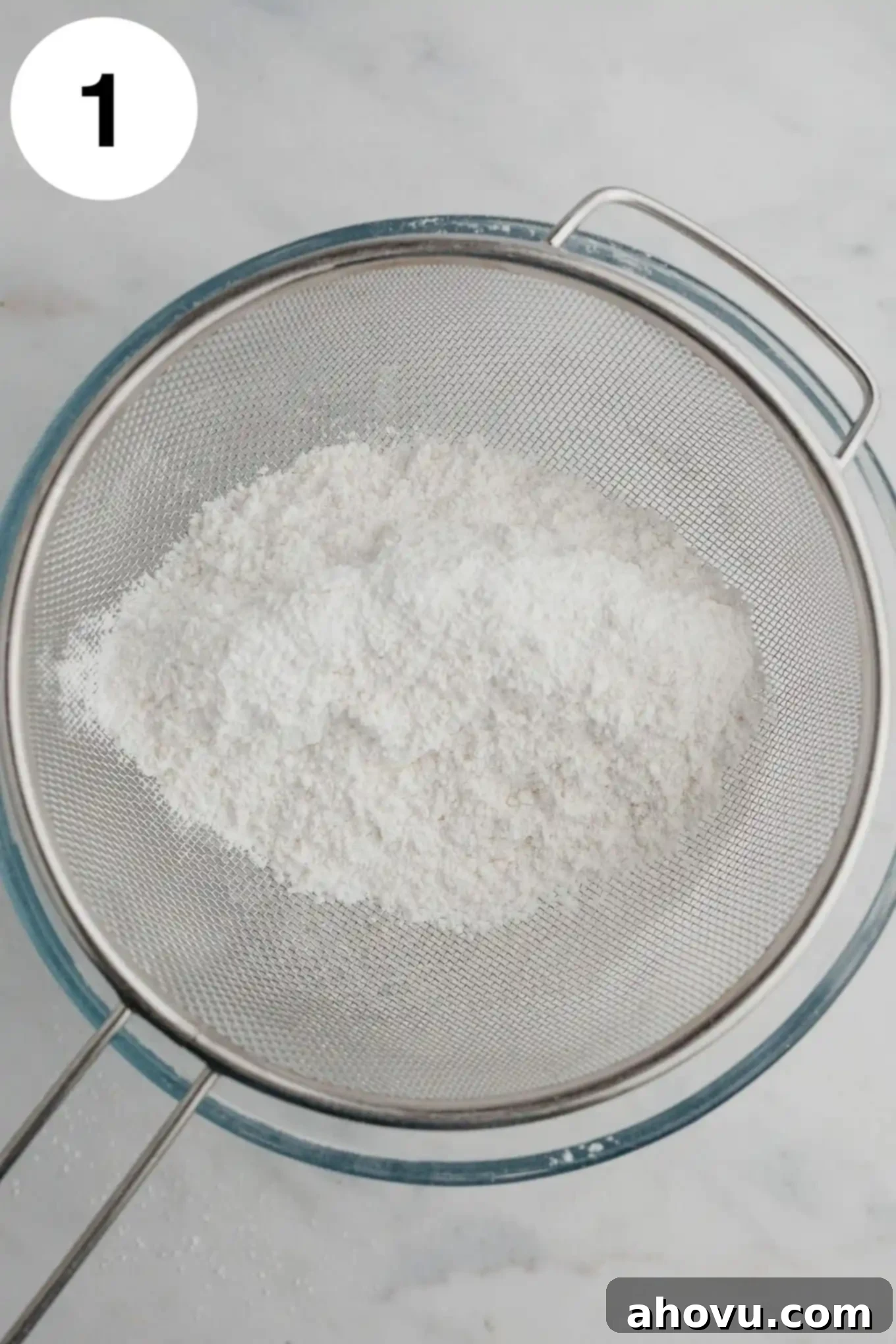 An overhead view of cake flour and powdered sugar being sifted through a fine mesh sieve into a glass mixing bowl.