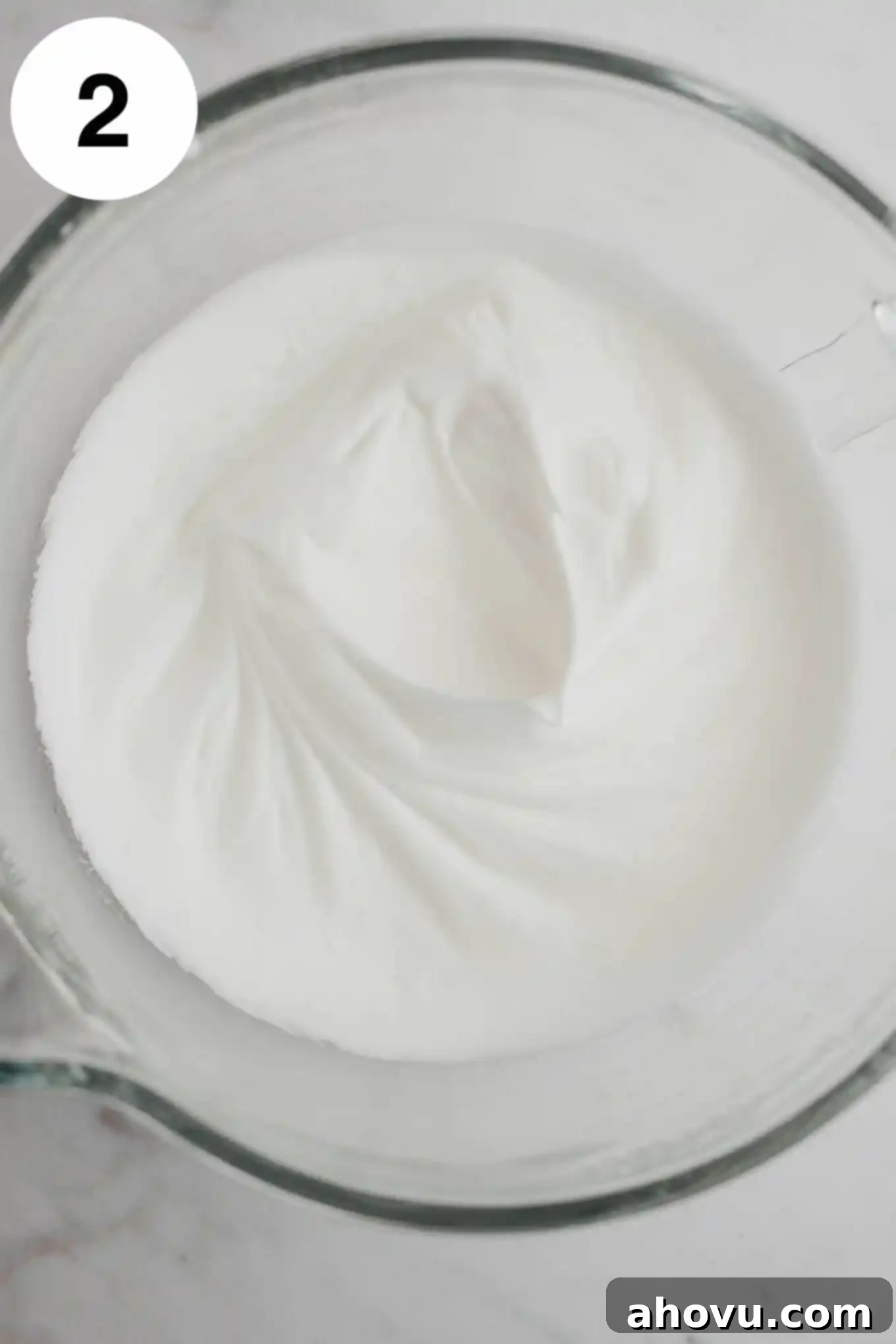 An overhead view of perfectly whipped egg whites forming stiff, glossy peaks in a clear glass mixing bowl, ready for the next step.