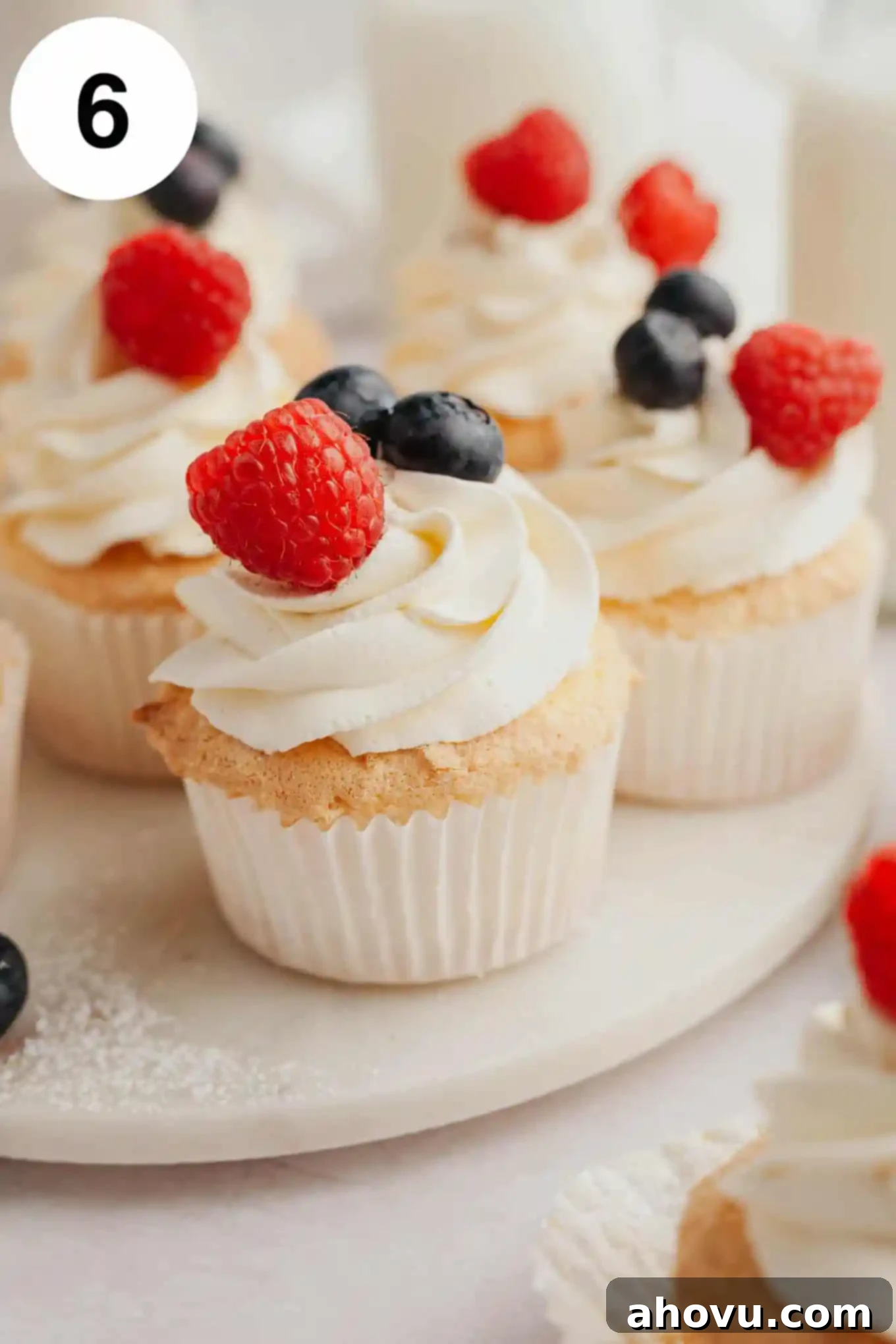 A close-up of several beautifully frosted angel food cupcakes, generously topped with whipped cream and a colorful assortment of fresh berries.