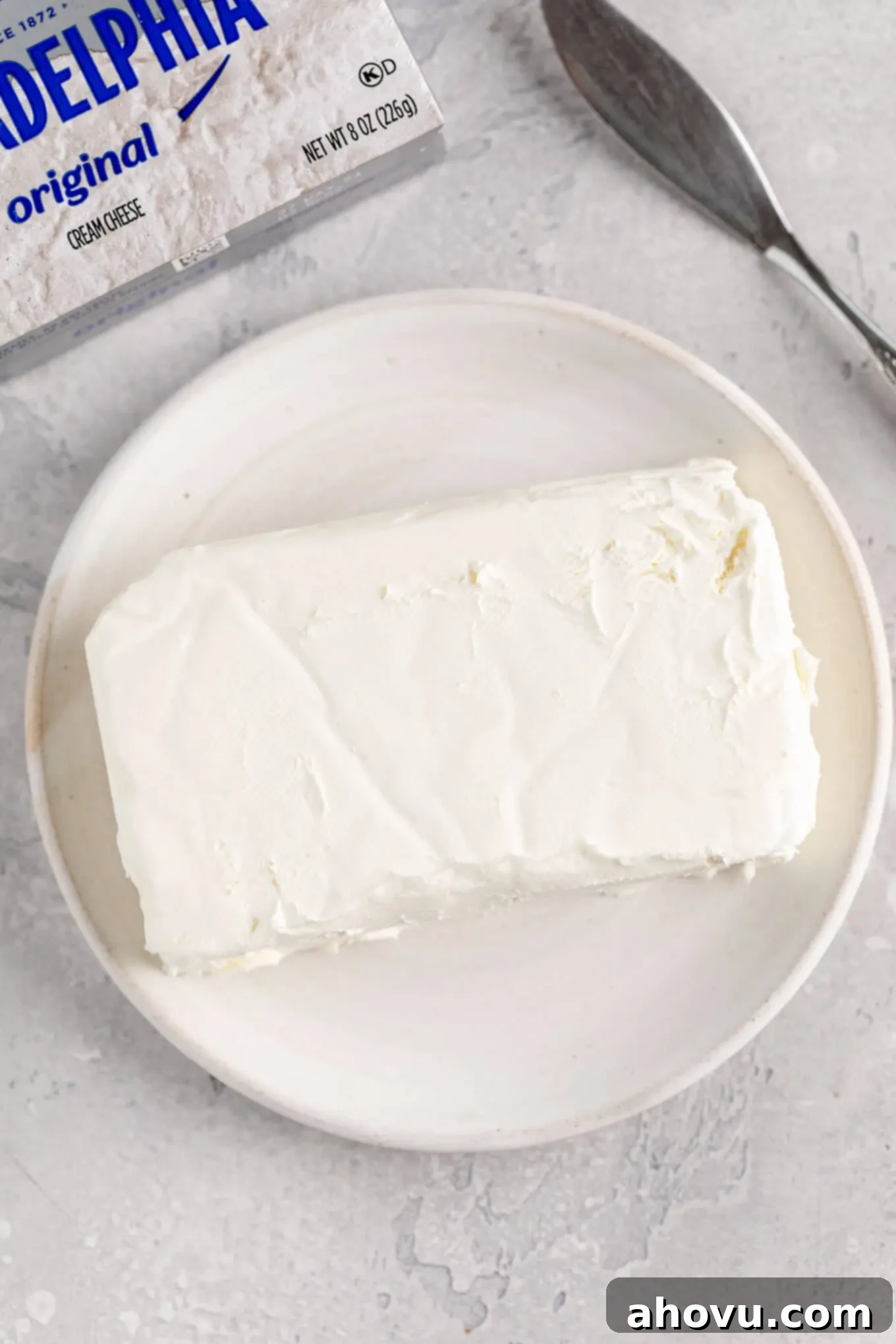 A block of cream cheese on a white plate. A box of Philadelphia cream cheese is sitting next to the plate, suggesting readiness for use in a recipe.
