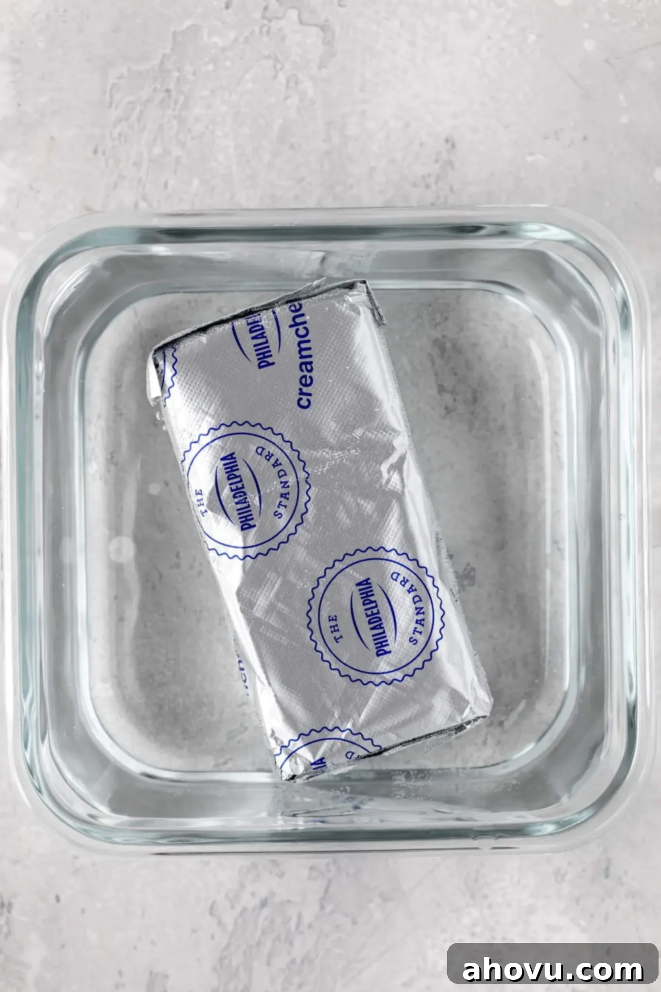 An overhead view of a sealed package of cream cheese nestled in a small container filled with warm water, demonstrating a rapid softening technique that maintains integrity.