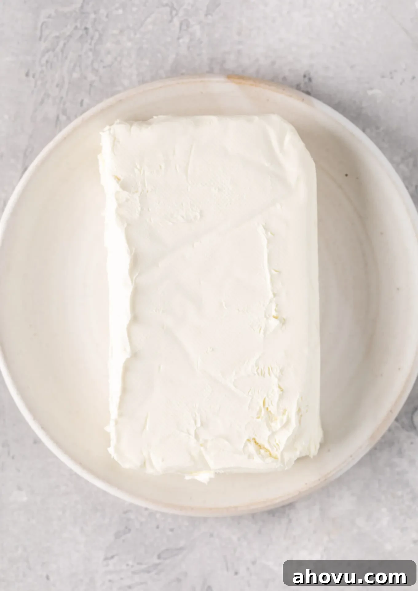 An overhead view of a block of cream cheese on a white plate, precisely positioned for microwave softening to ensure even heating.