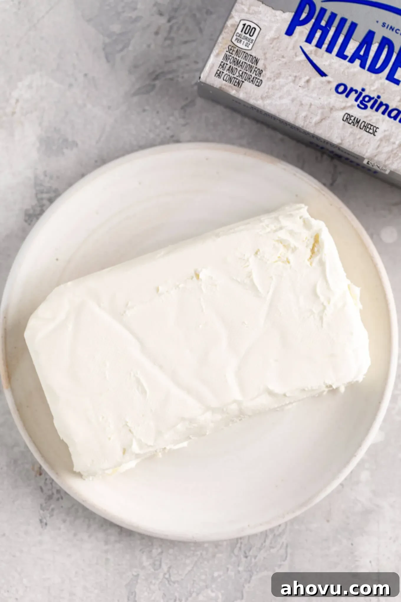 A block of perfectly softened cream cheese on a white plate, with its original Philadelphia cream cheese box nearby, indicating readiness for baking.