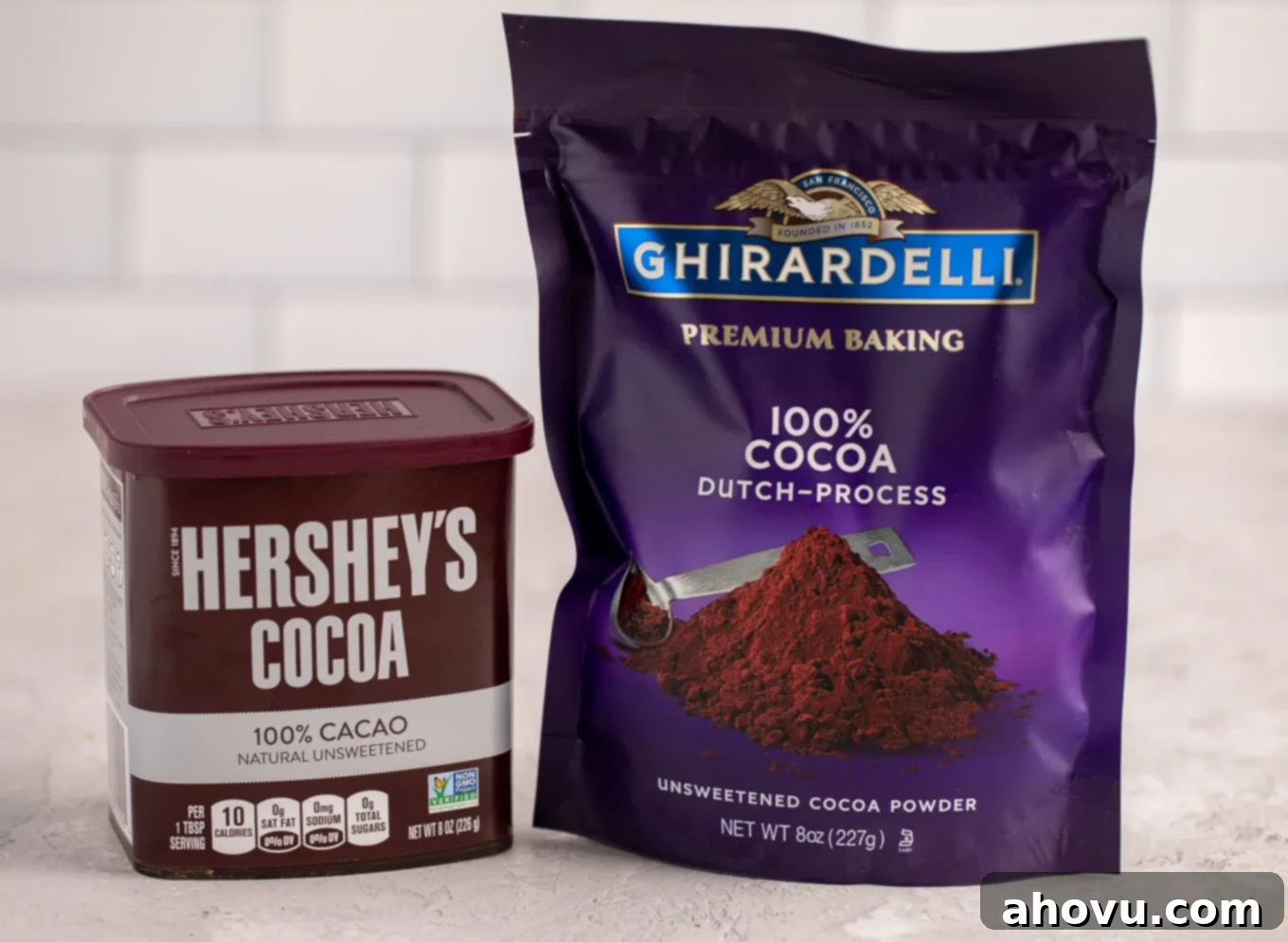 A container of Hershey's unsweetened cocoa powder and a package of Ghirardelli Dutch process cocoa powder sitting next to each other on a gray surface.