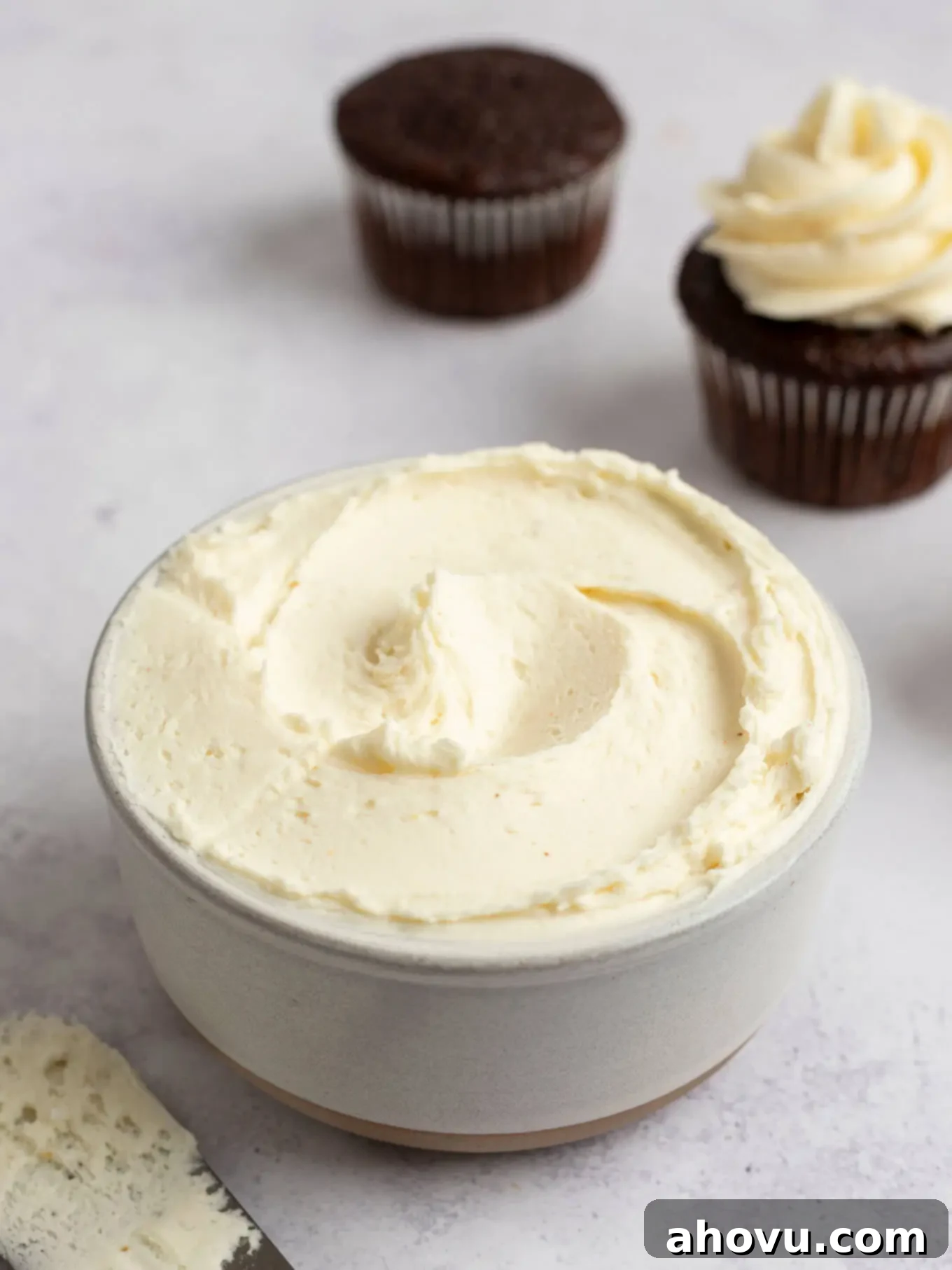 A pristine white bowl overflowing with velvety brown butter frosting, positioned artfully near two exquisitely frosted chocolate cupcakes.