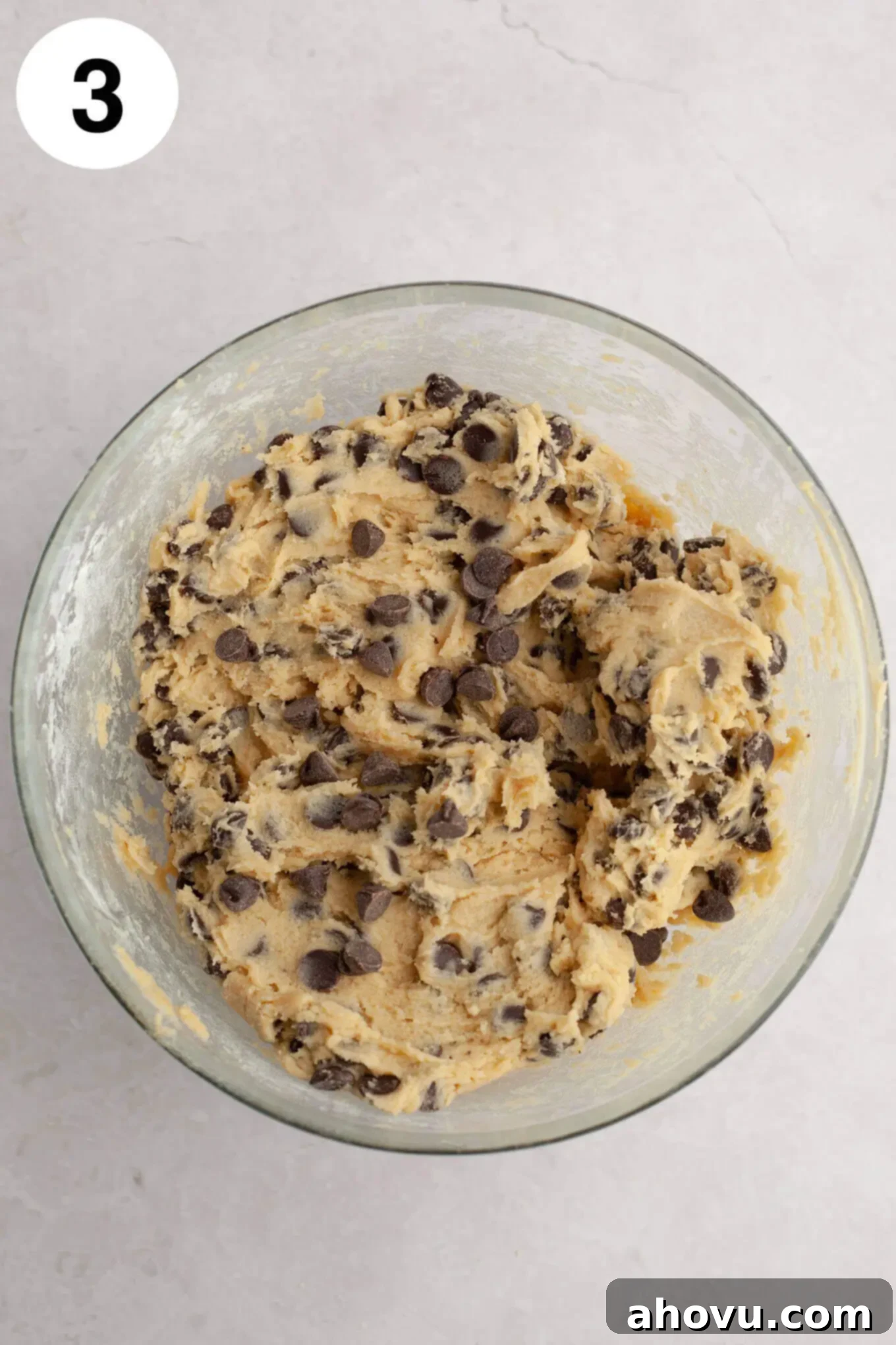 An overhead view of rich chocolate chip cream cheese cookie dough in a glass mixing bowl, speckled with chocolate chips.