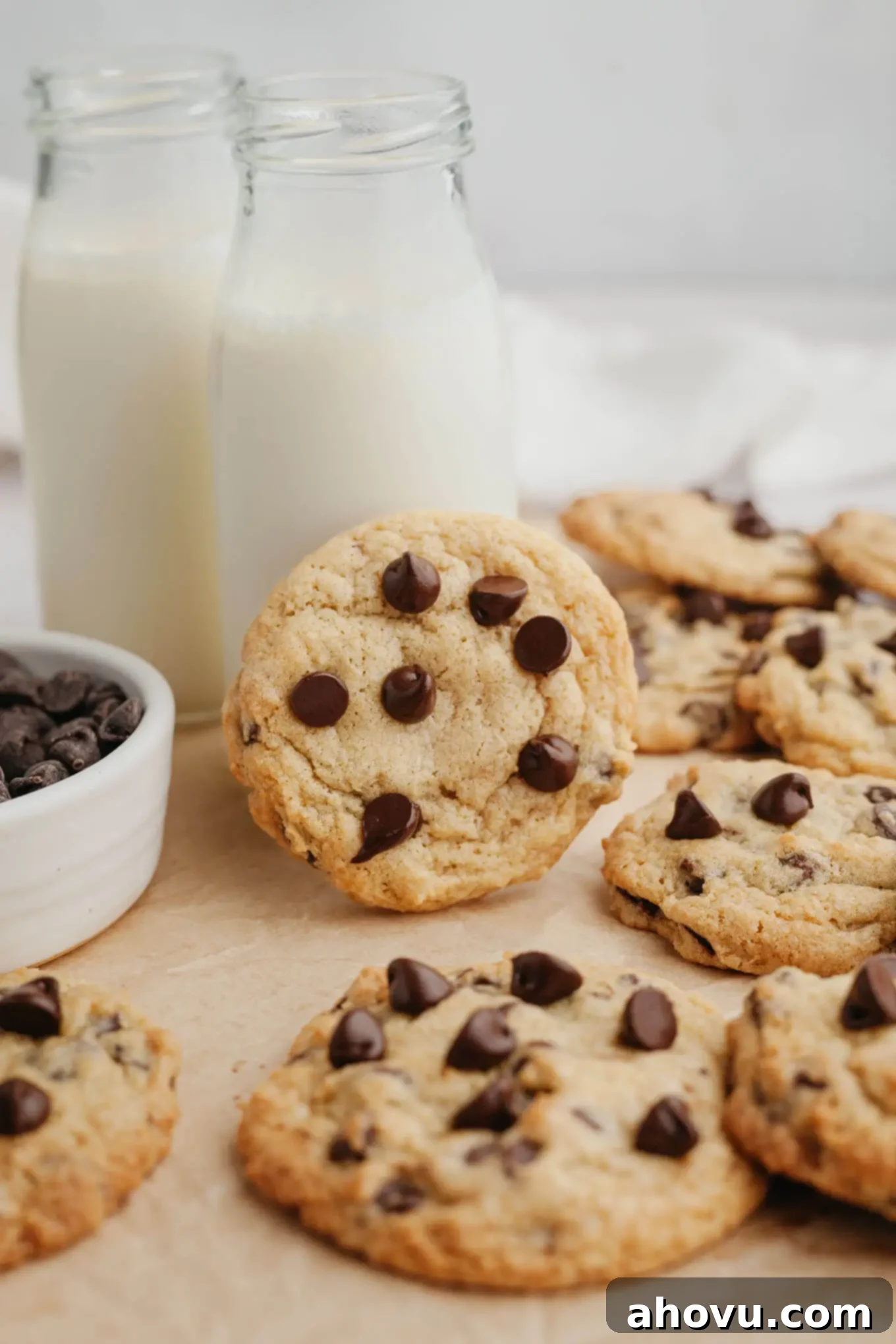 Two glass milk jugs surrounded by warm, freshly baked cream cheese chocolate chip cookies. One cookie leans invitingly against a milk jug, perfect for dipping.