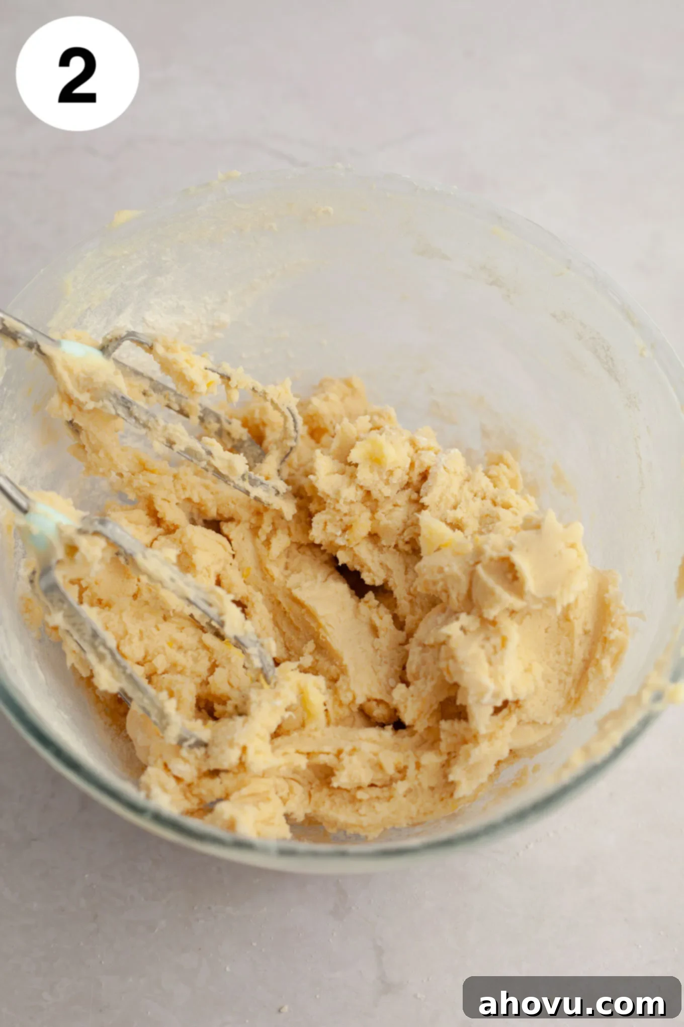 An overhead view of smooth, pale yellow lemon cookie dough in a glass mixing bowl, with electric beaters on the side, indicating it's perfectly mixed.