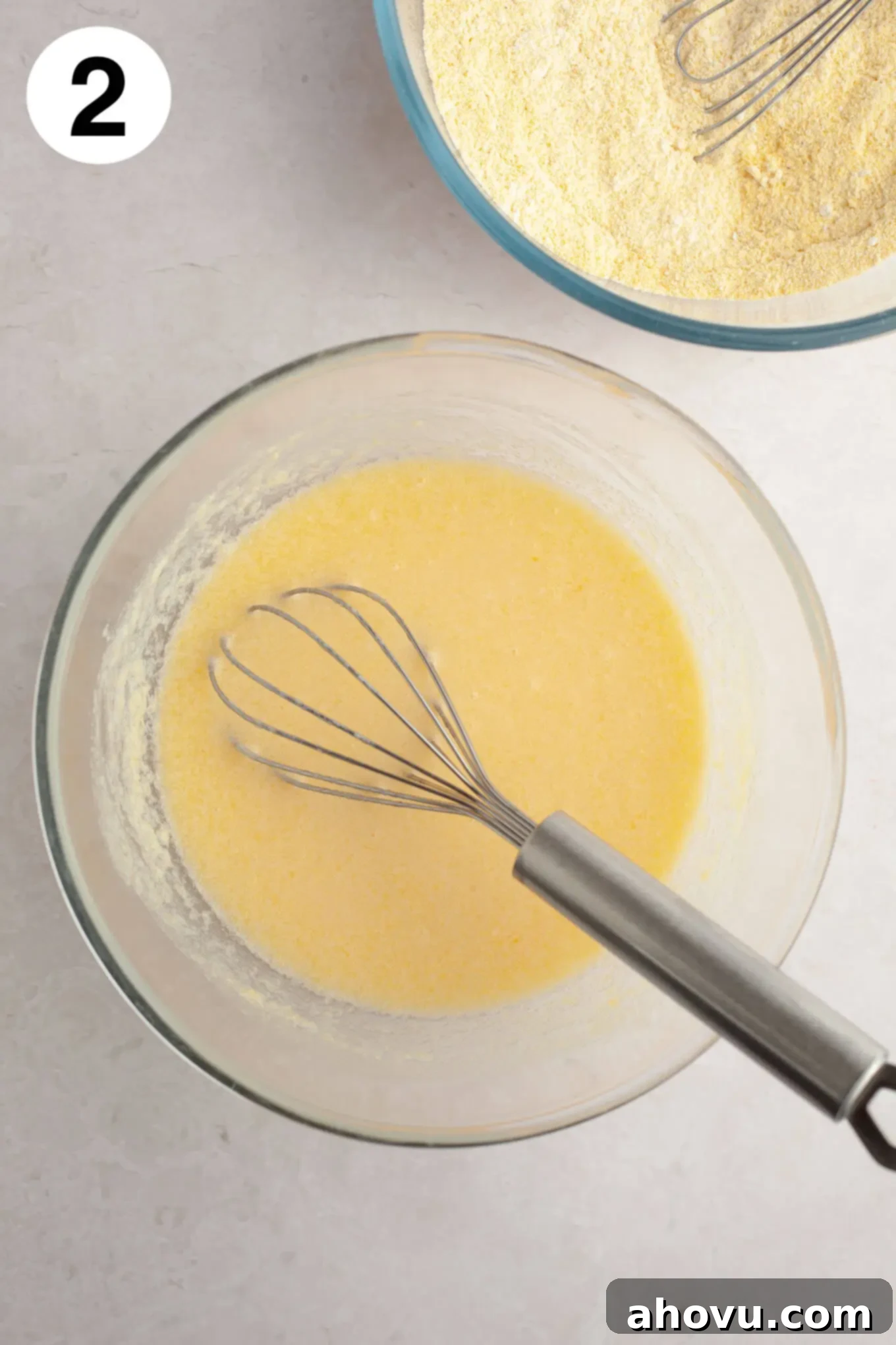 An overhead view of the wet ingredients for cornbread muffins in a glass mixing bowl with a wire whisk. 