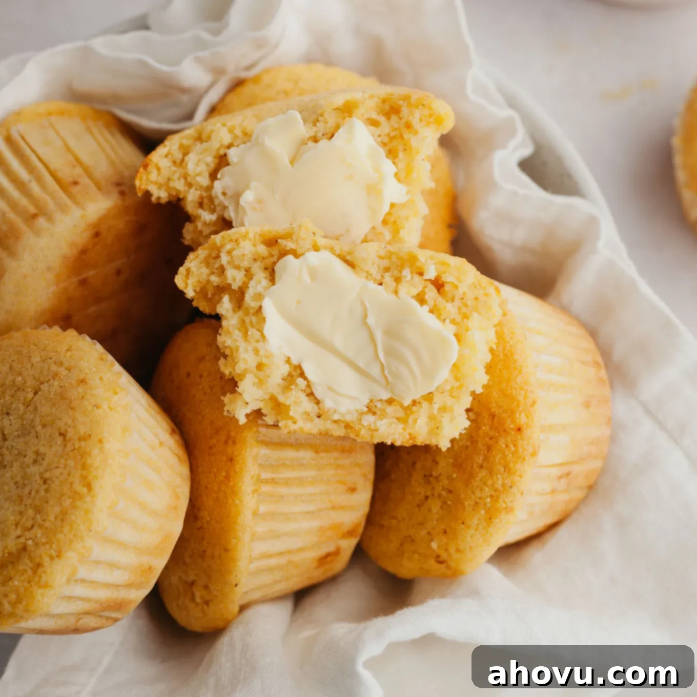 A pile of cornbread muffins in a towel-lined serving bowl. The top muffin has been split in half and slathered with butter. 