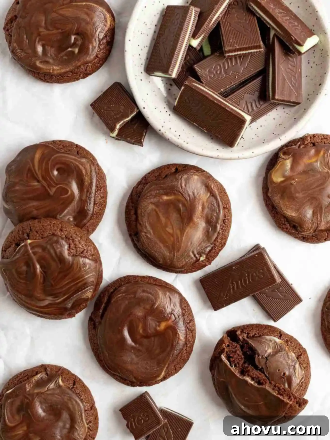 An overhead view of several finished Andes mint cookies, beautifully spread with melted mint icing, arranged on parchment paper alongside a bowl of remaining Andes mints.