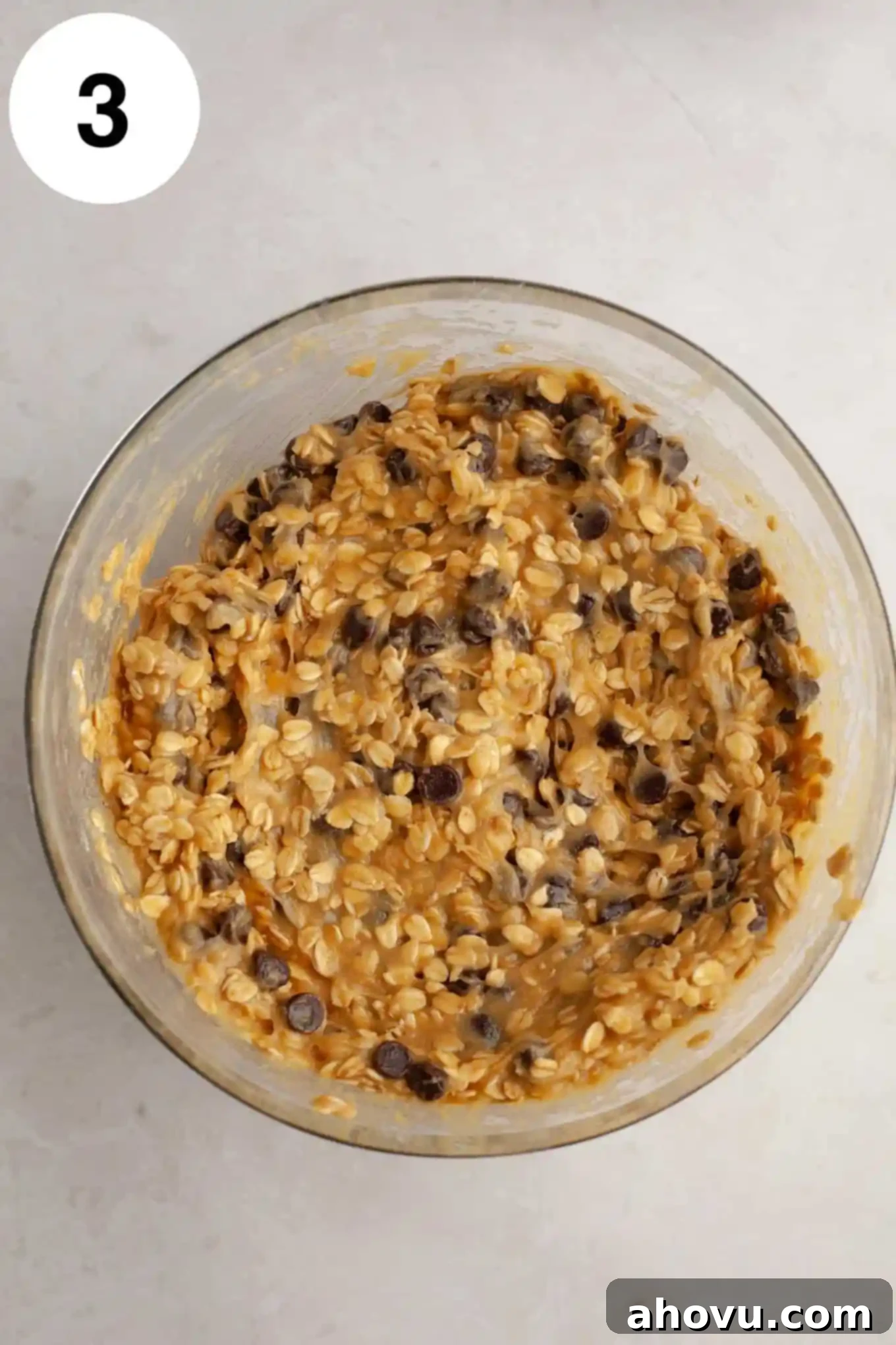 An overhead view of oatmeal chocolate chip cookie dough in a clear glass mixing bowl, ready for baking.