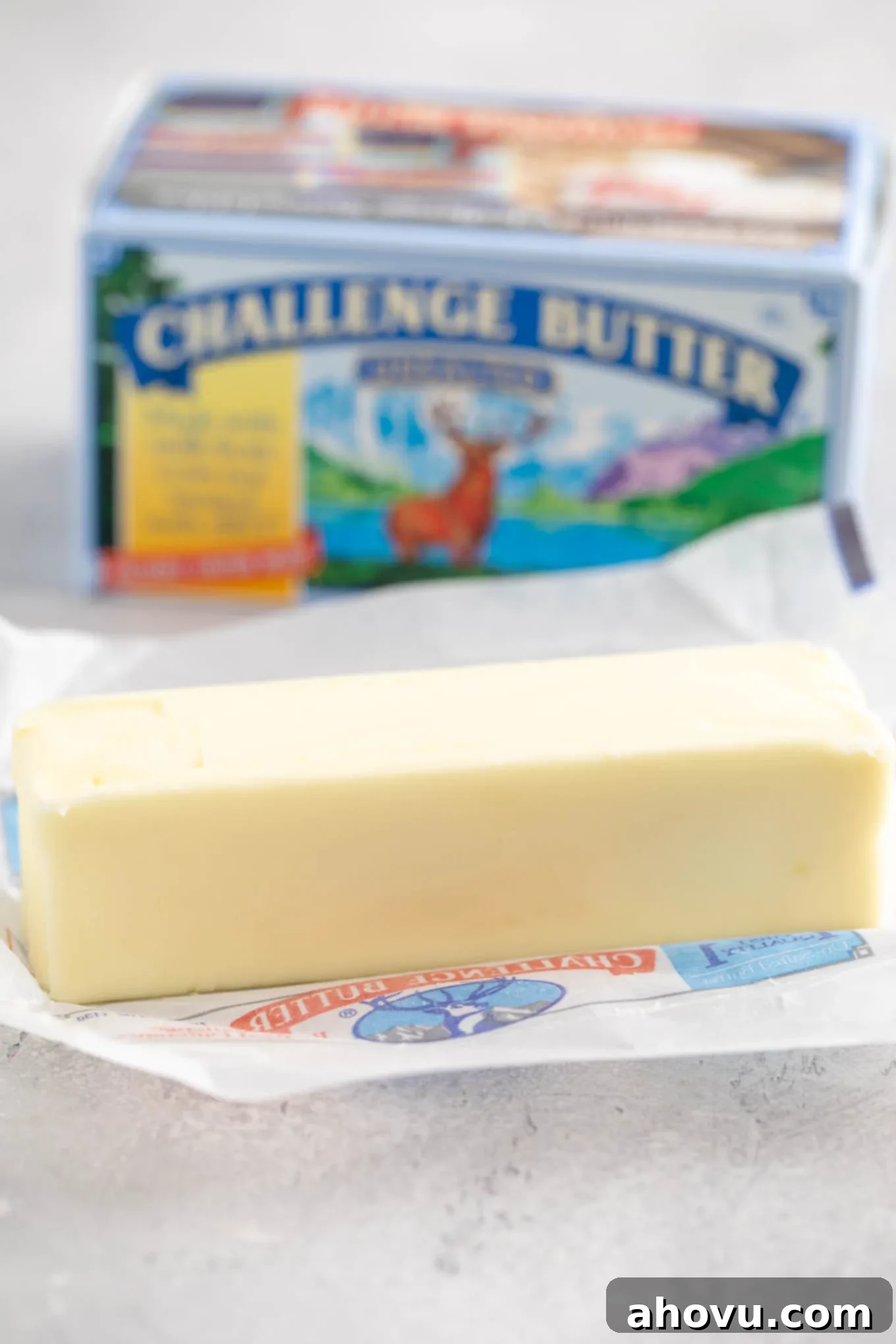 An opened stick of unsalted butter sitting on top of the wrapper. A box of butter is sitting in the background.