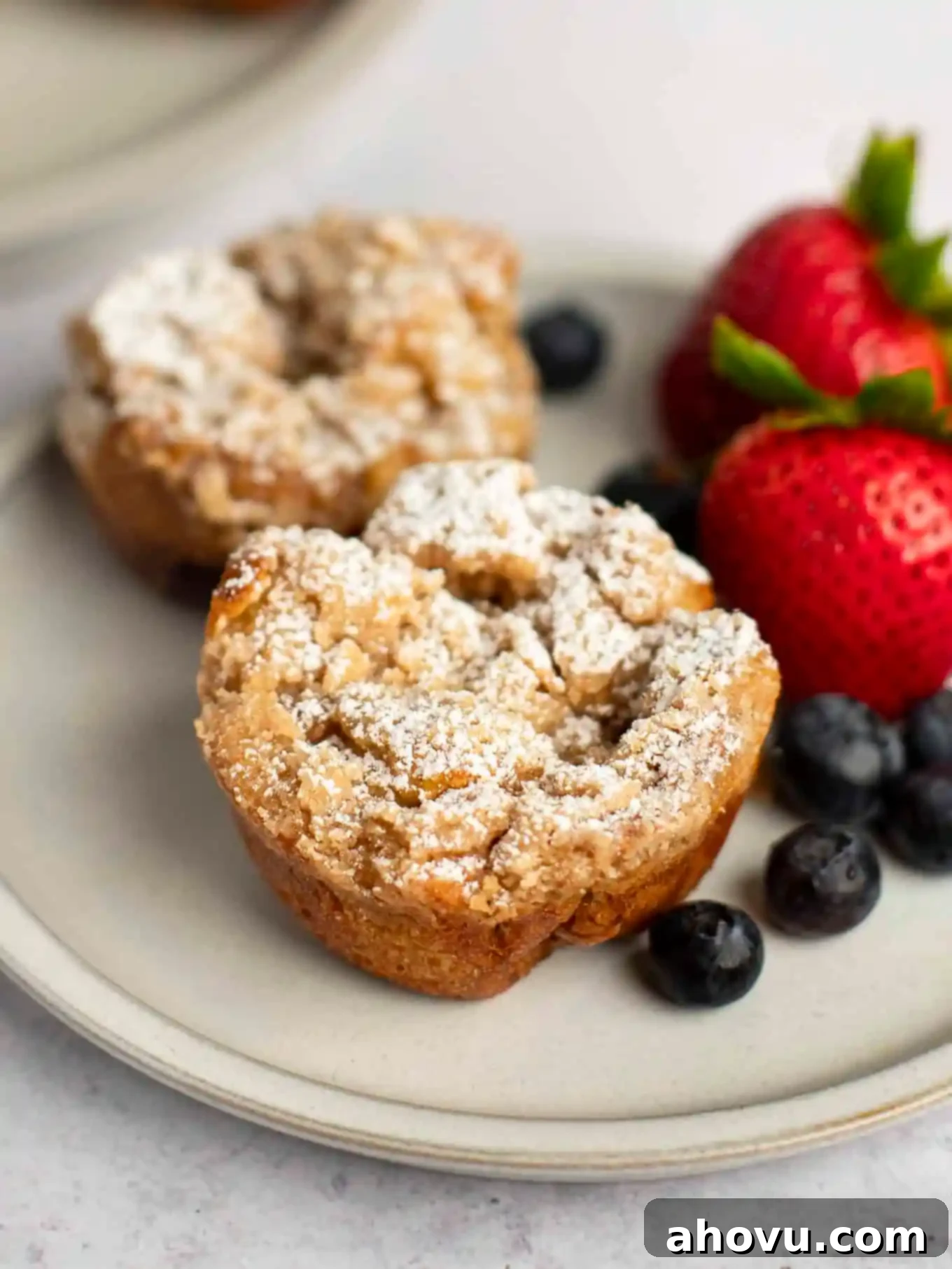 Two French toast muffins dusted with powdered sugar sitting on a plate next to fresh fruit.