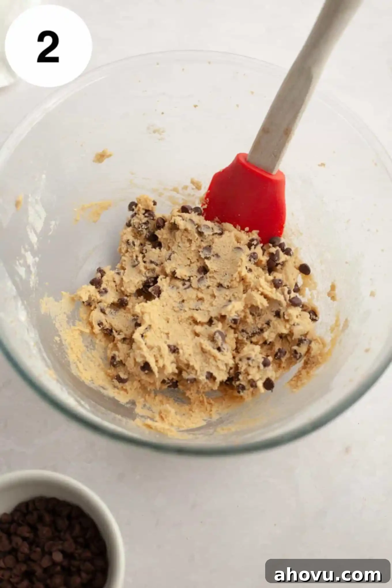 An overhead view of edible chocolate chip cookie dough in a glass mixing bowl, with a rubber spatula. 