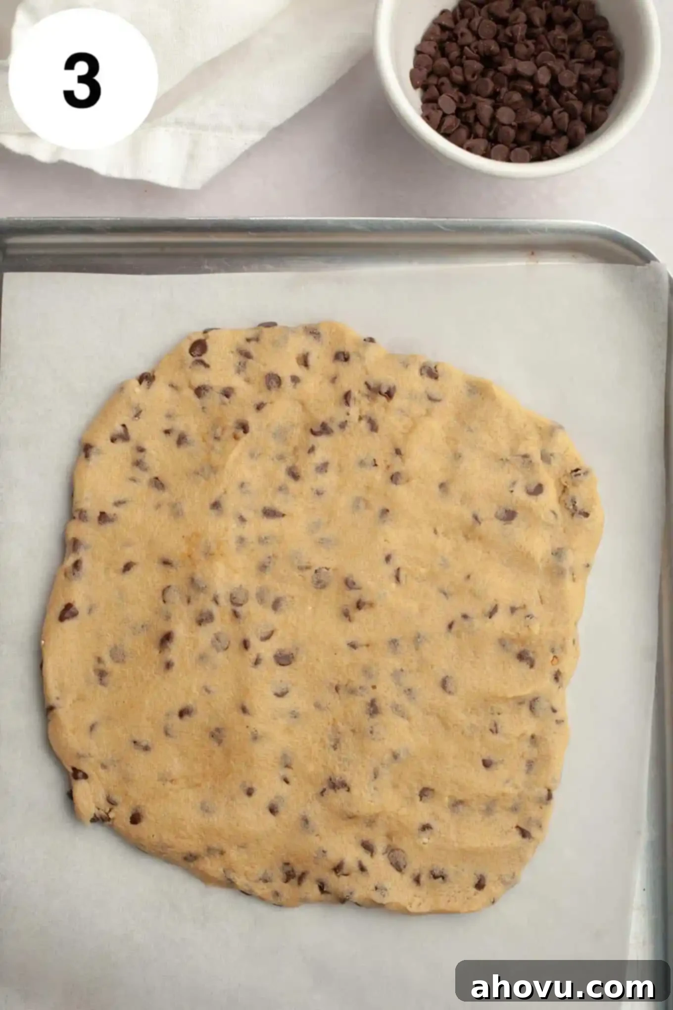 An overhead view of edible chocolate chip cookie dough spread on a parchment paper-lined baking sheet. 