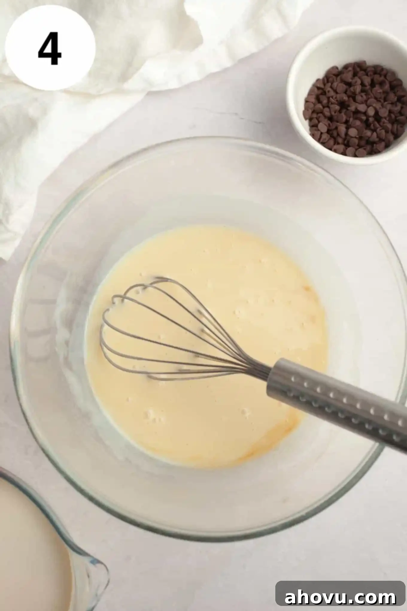 An overhead view of a sweetened condensed milk mixture in a glass mixing bowl, with a whisk