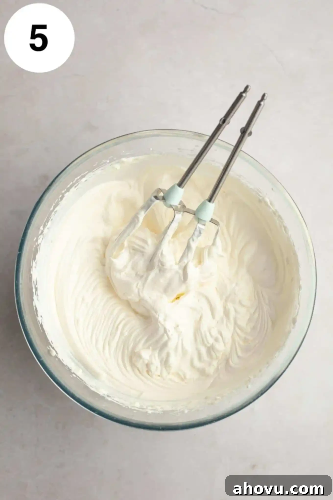 An overhead view of whipped cream in a glass mixing bowl, with two electric beaters. 