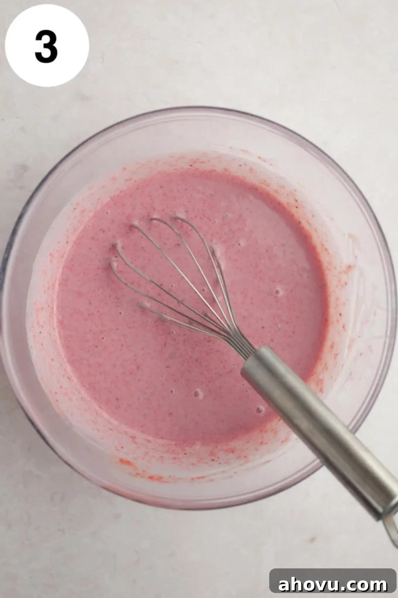 An overhead shot of sweetened condensed milk and the vibrant, cooled strawberry mixture being combined in a clear glass mixing bowl, showcasing the rich color.