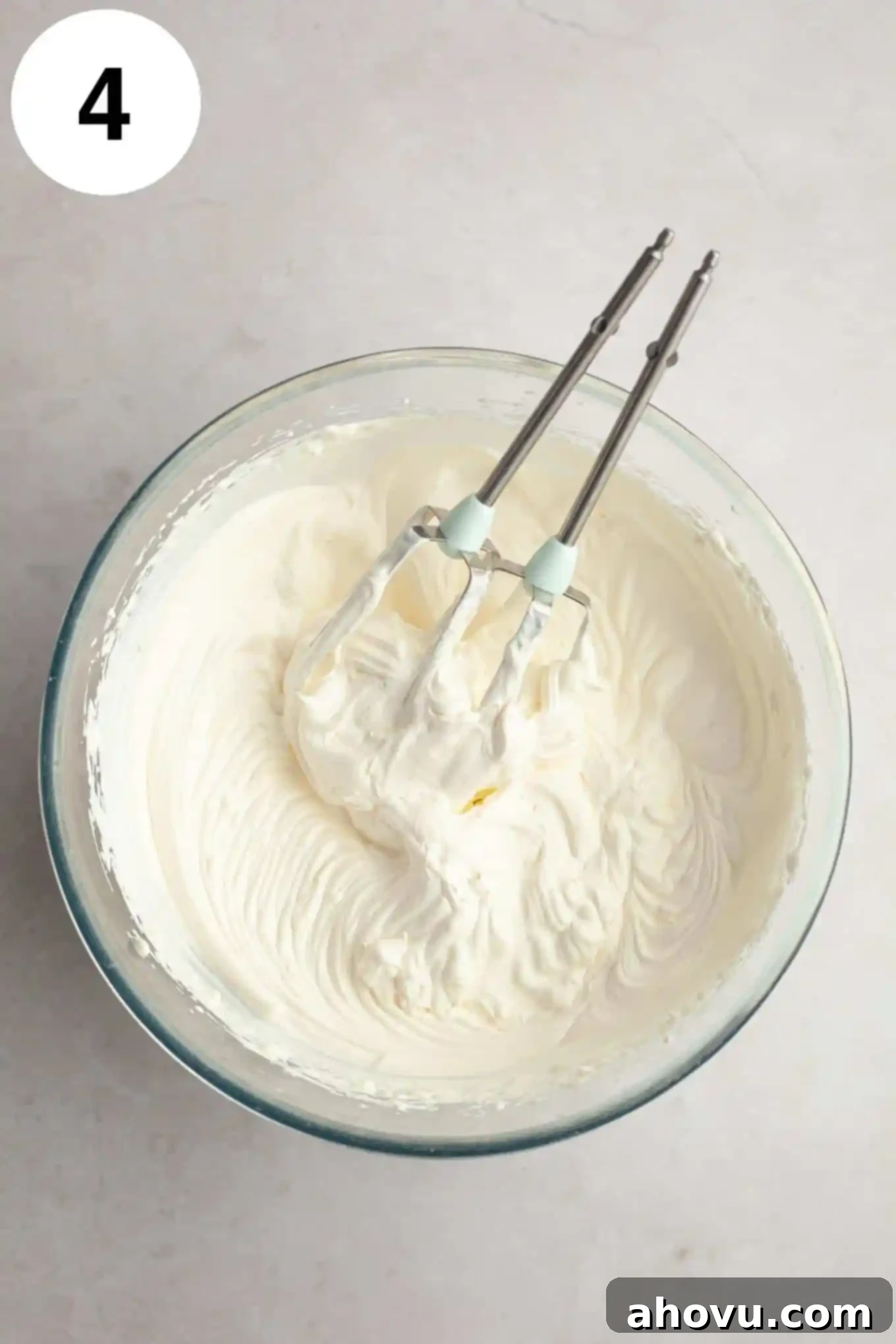 An overhead view of fluffy, stiff-peaked whipped cream in a glass mixing bowl, with two electric beaters resting beside it, indicating readiness.
