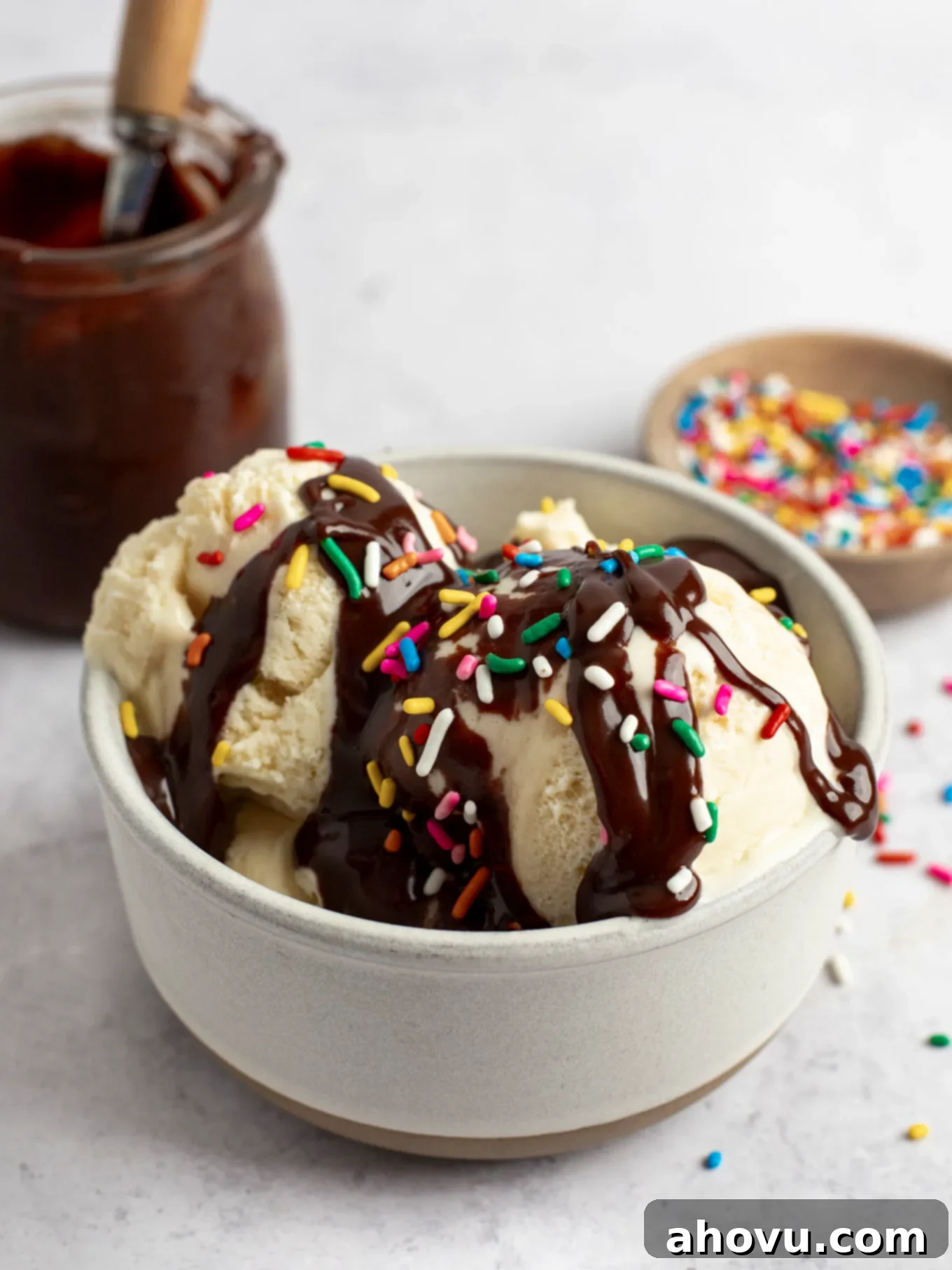 A dish of vanilla ice cream generously topped with dark hot fudge sauce and colorful rainbow sprinkles, inviting a sweet indulgence.